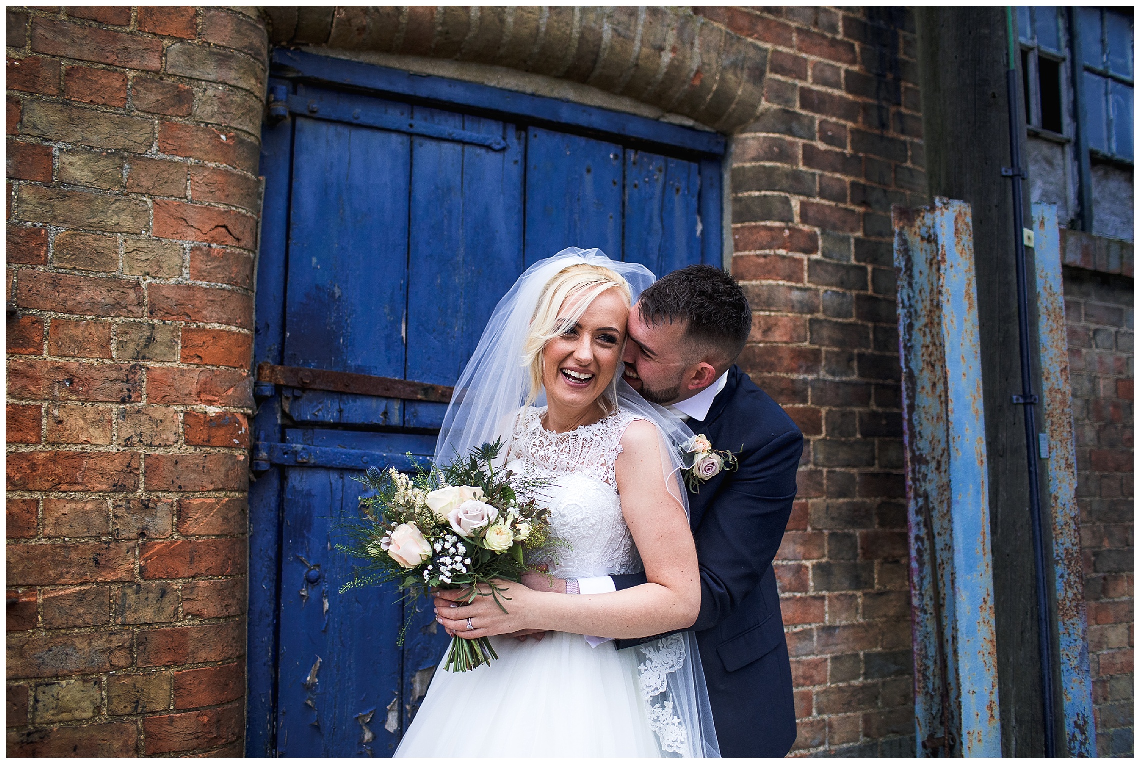 groom standing behind bride and nuzzling her neck, stood in front of building with a blue door at bassmead manor barns