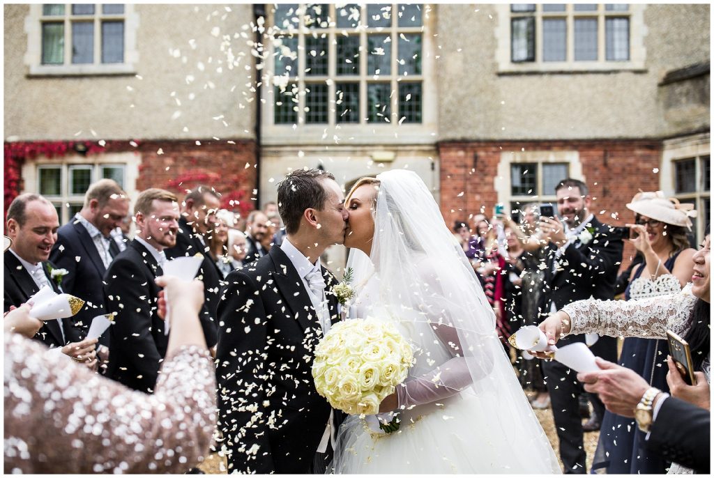 Bride and groom kiss as wedding guests throw confetti over them, outside at plum park. 