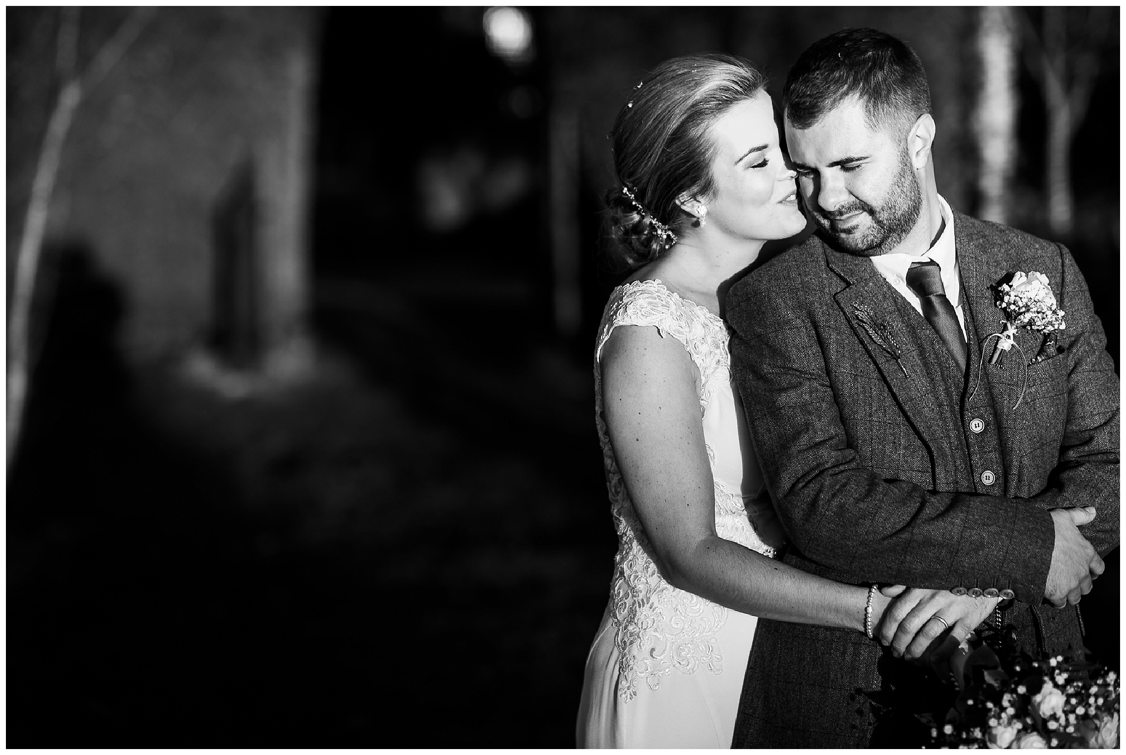 Bride standing behind groom and kissing him on cheek