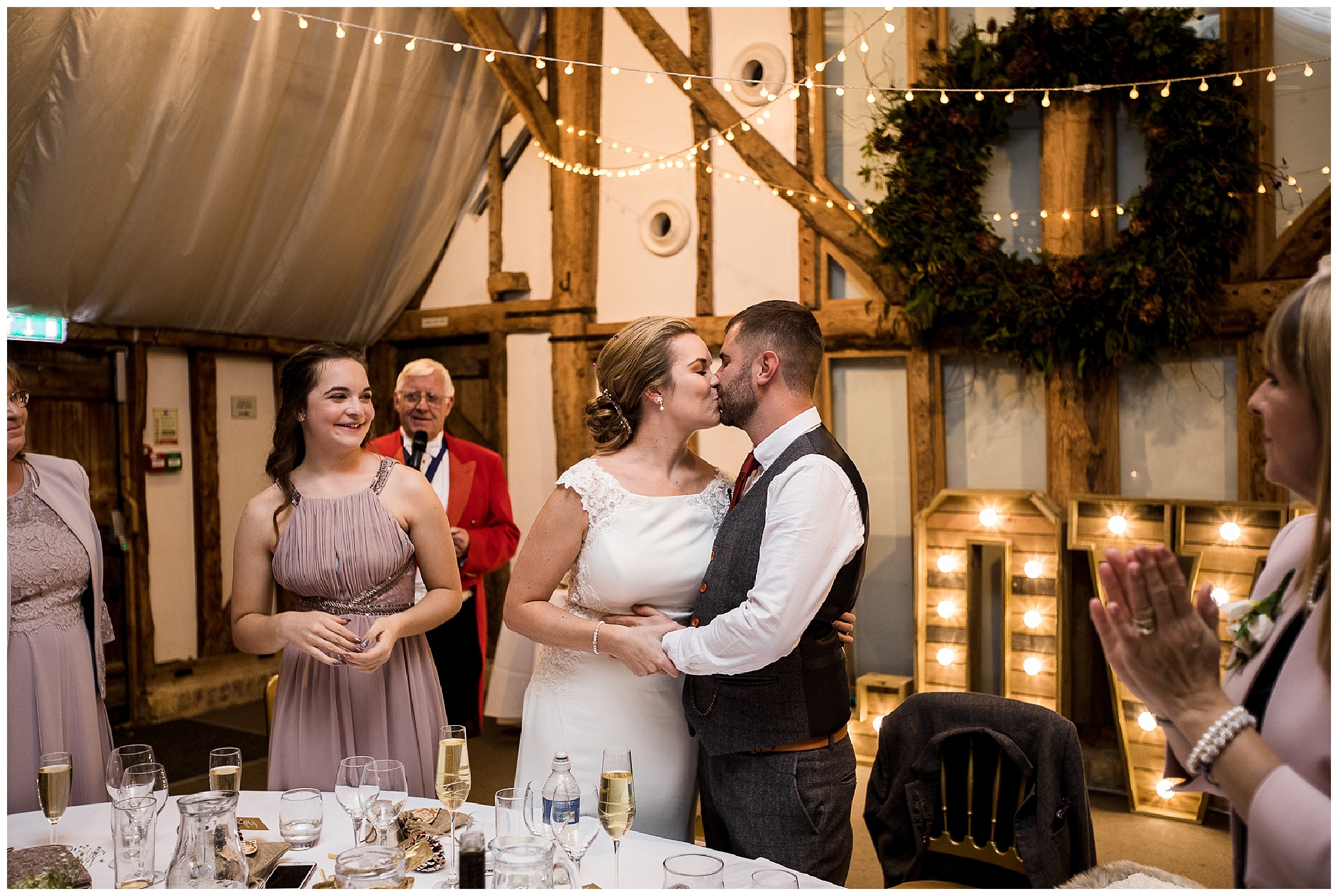 bride and groom kiss at top table with family onlooking and smiling after speeches