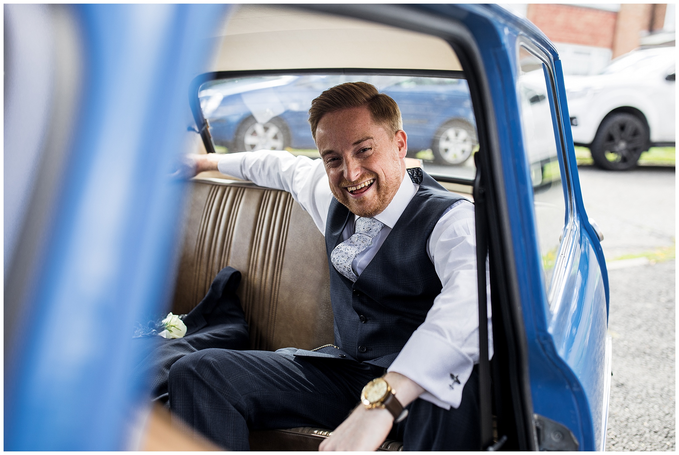 Groom sat in the back of a blue mini in a grey suit laughing
