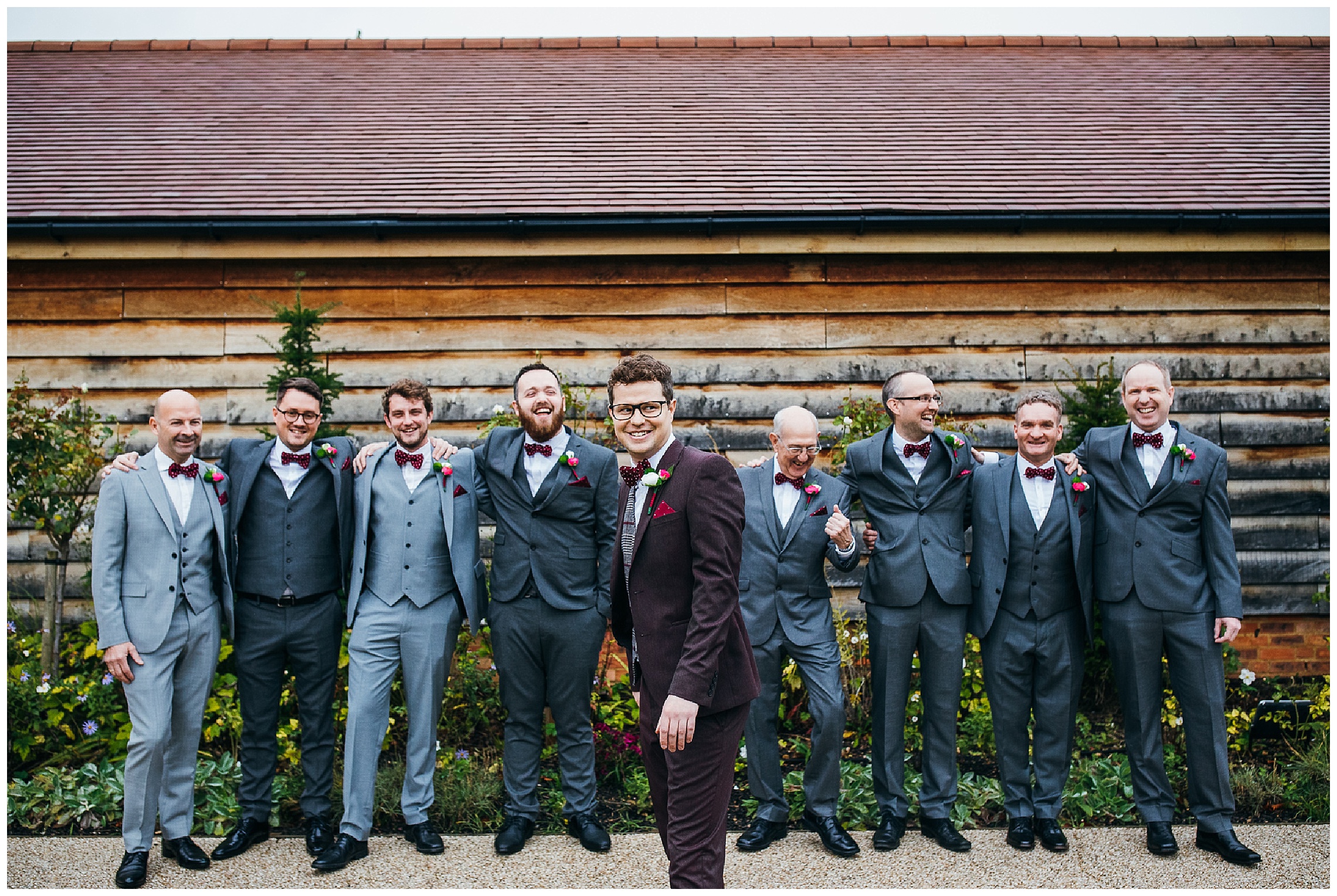 Groom in burgundy suit amongst groomsmen in grey suits, outside Bassmead barn wedding venue