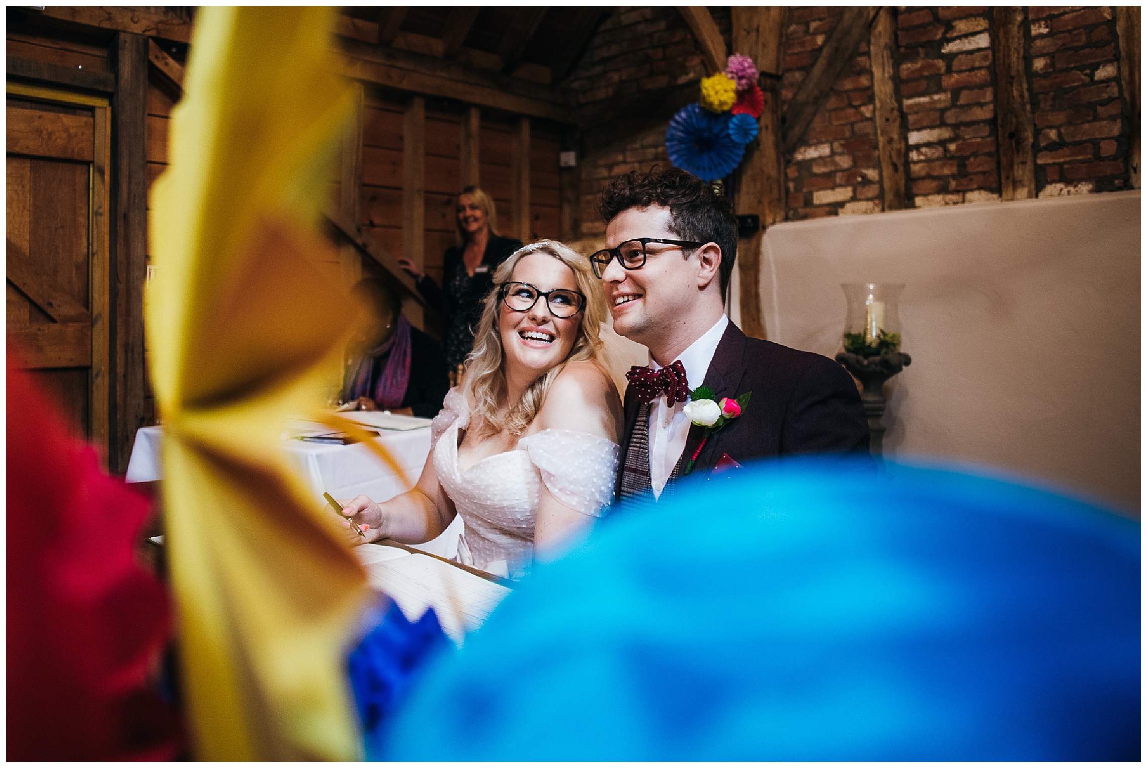 bride looks over at groom as they sign the register surrounded by colourful decorations