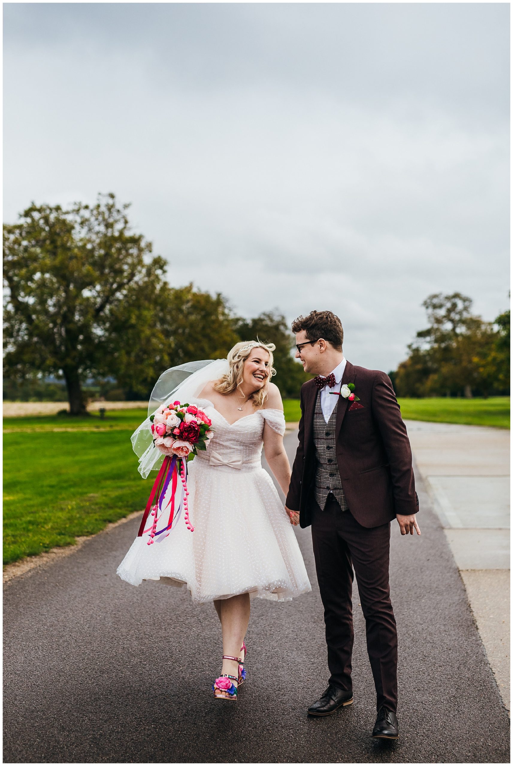 Bride in short dress and colourful shoes smiles at groom in burgundy suit and bow tie