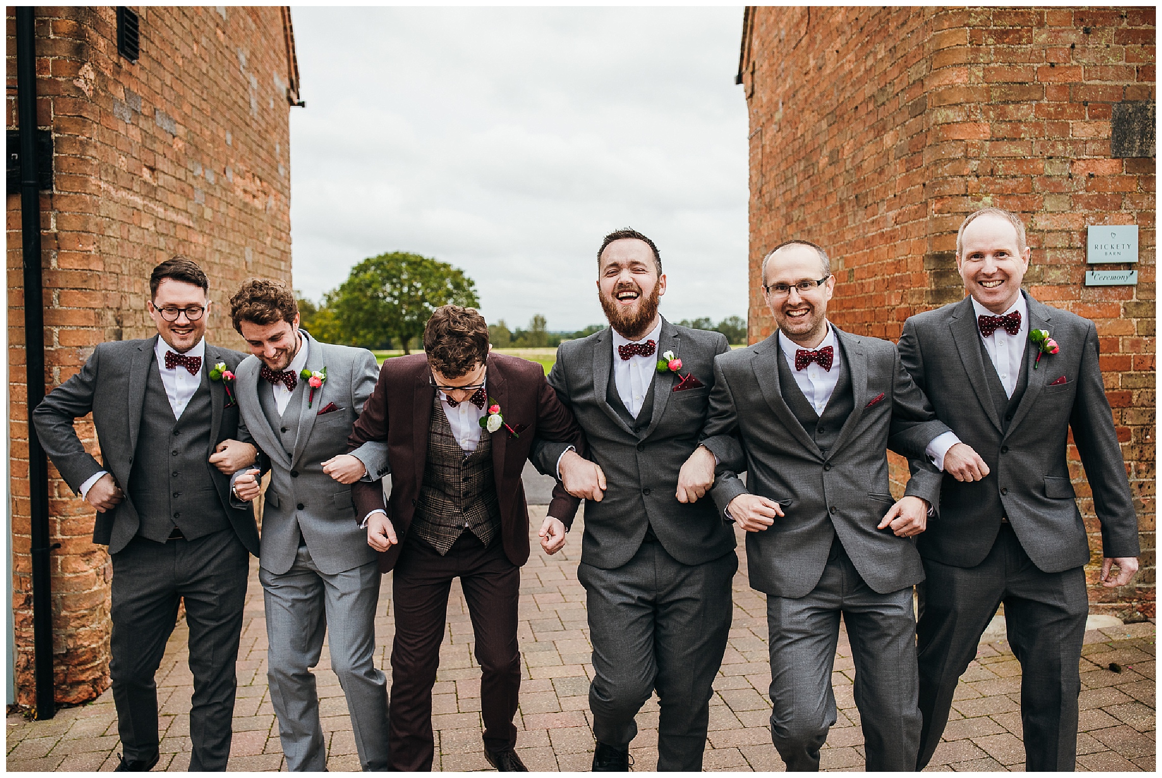 Groomsmen in grey and burgundy suits walk together between brick buildings at bassmead manor barns