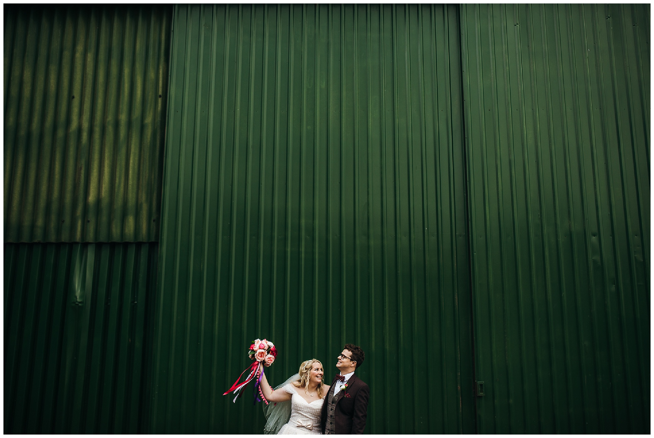 bride holds pink flowers in air next to groom in front of industrial barn at bassmead manor