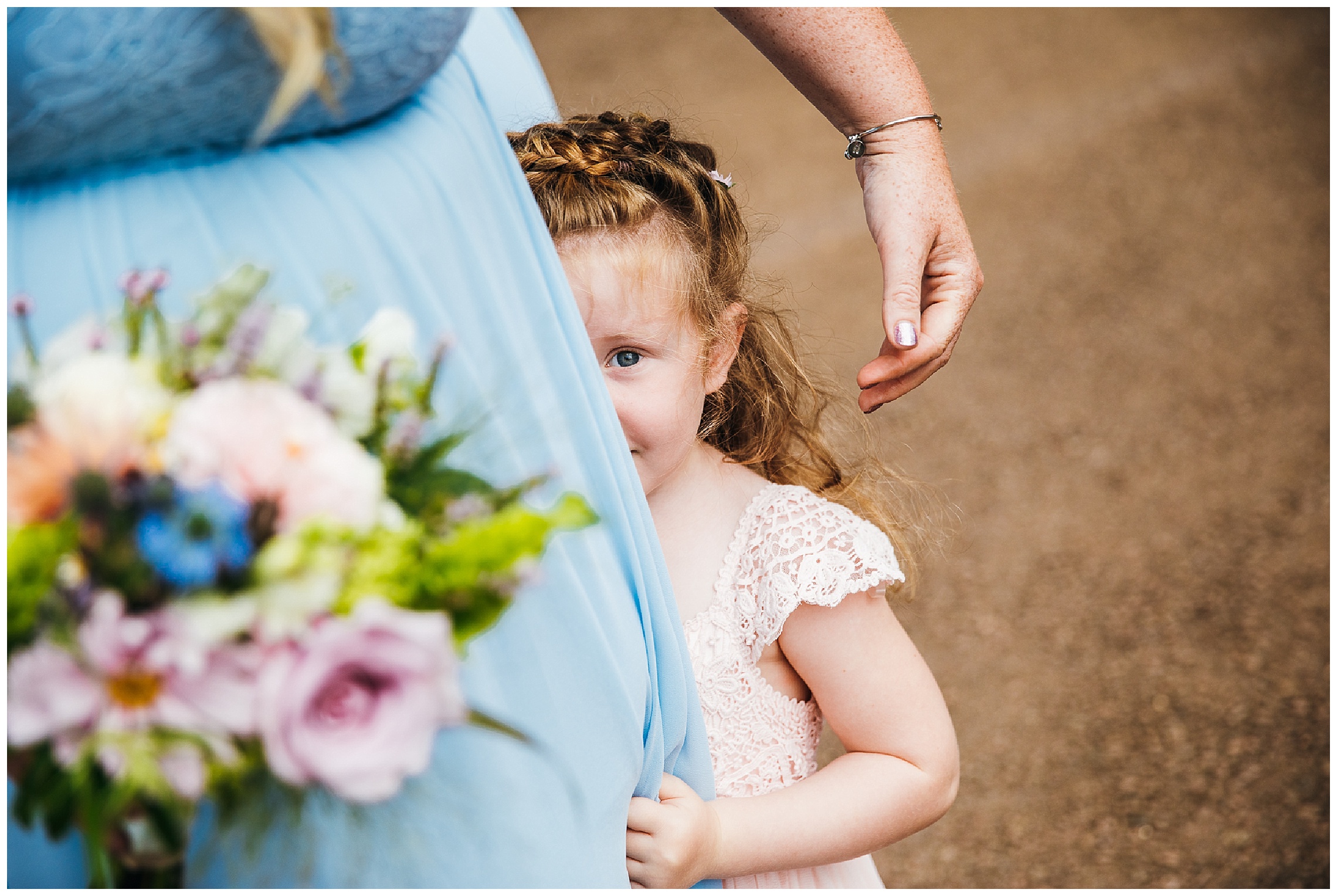 little flower girl in pink dress peers out from behind her mums legs