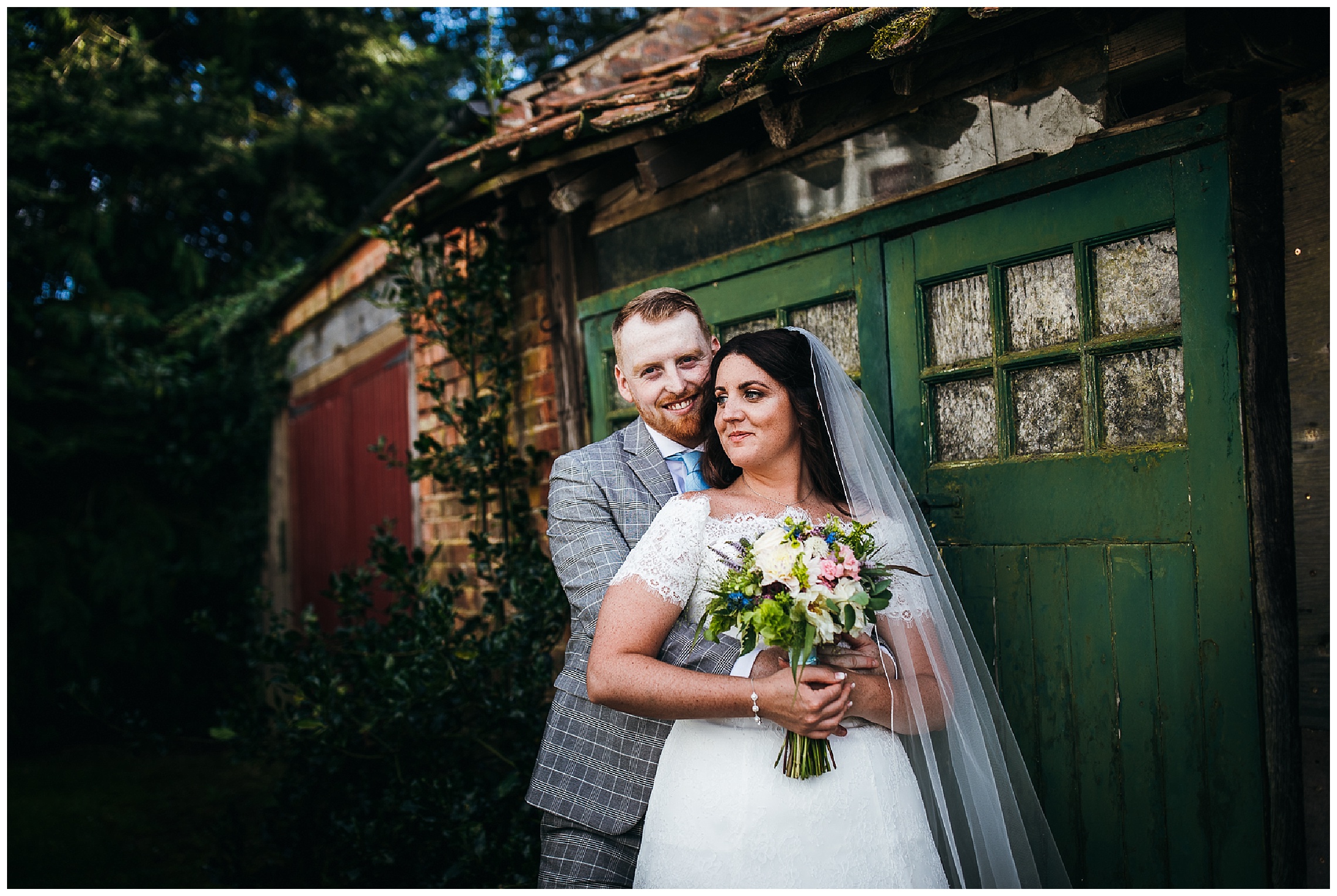 bride and groom stand in front of red and green rustic doors and smile together at hunsbury hill wedding venue