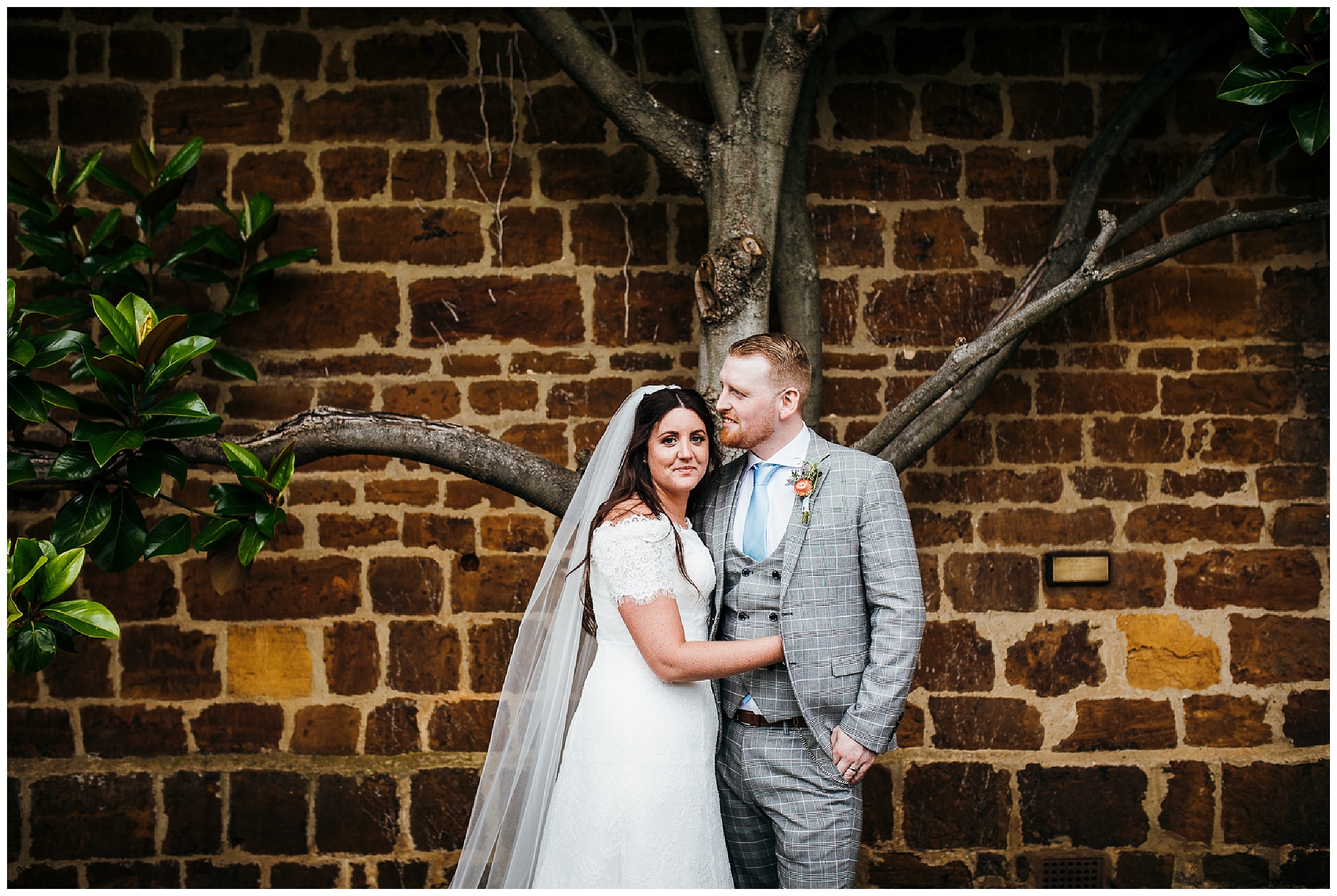 bride and groom stand together in front of tree and brick wall