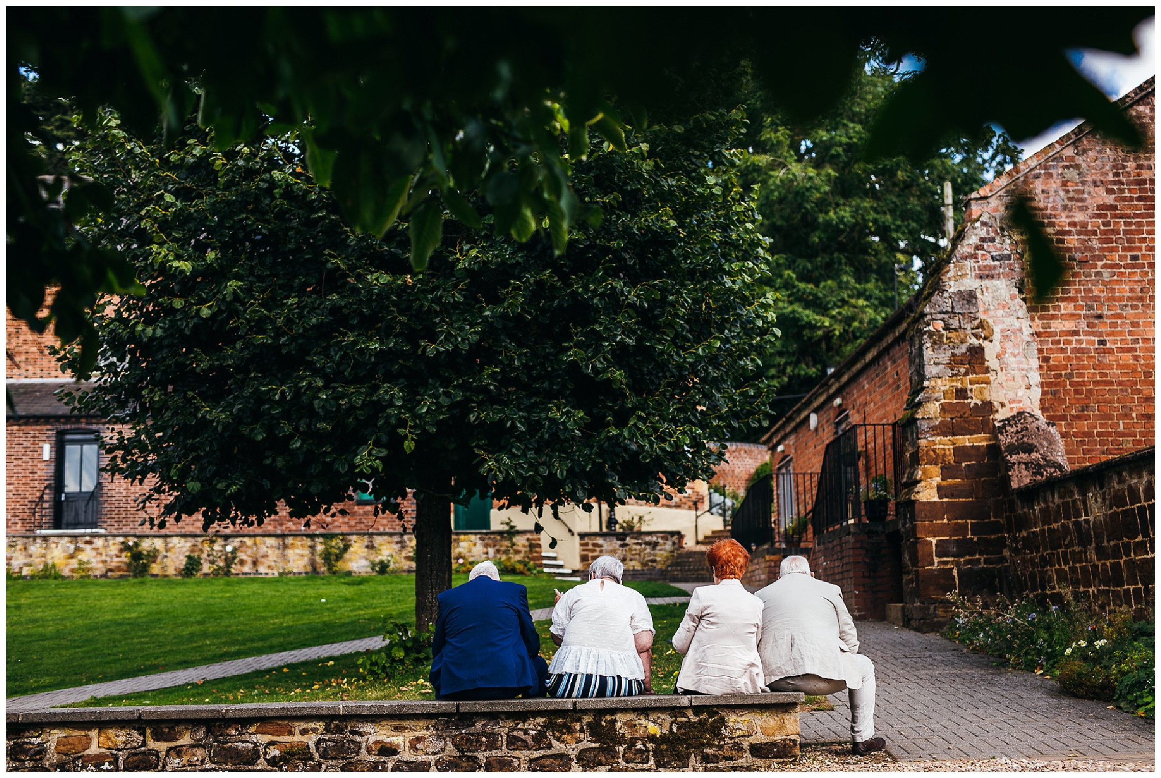wedding guests sit on brick wall and talk to one another on sunny day