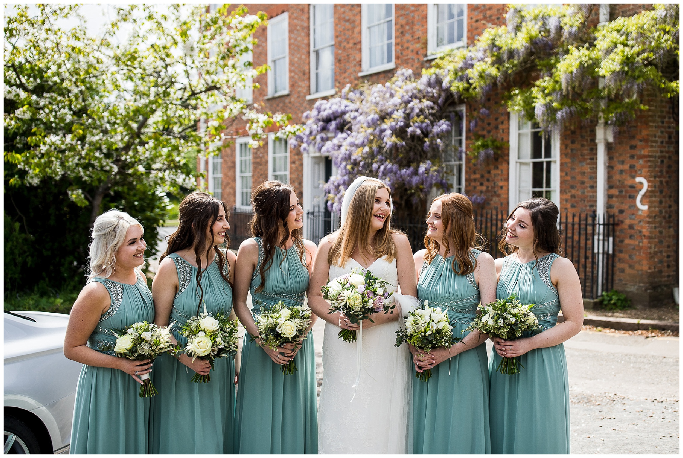 bride and bridesmaids hold white flower bouquets outside church with wisteria on a sunny day
