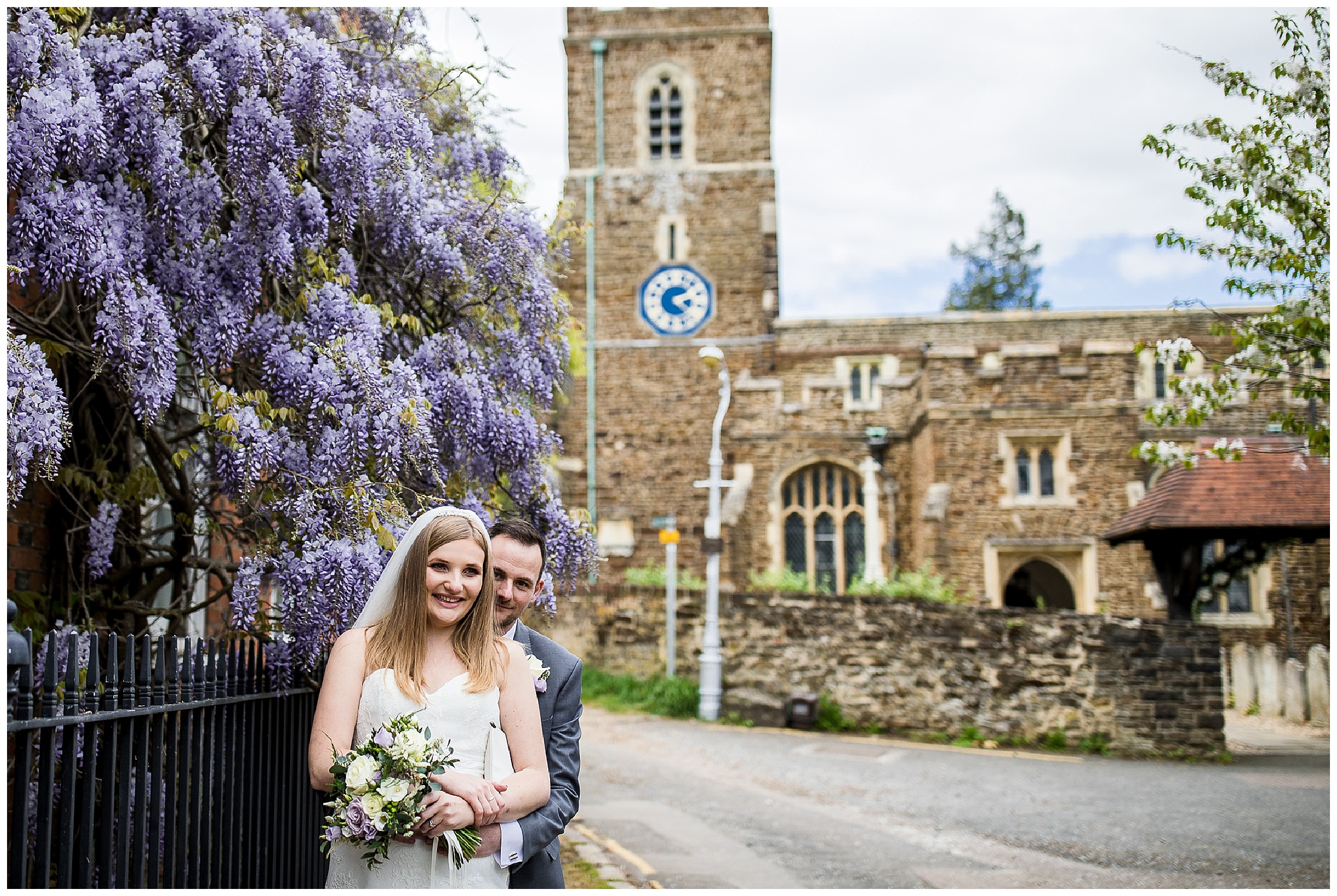 Bride and groom stand together in front of wisteria at Ampthill church