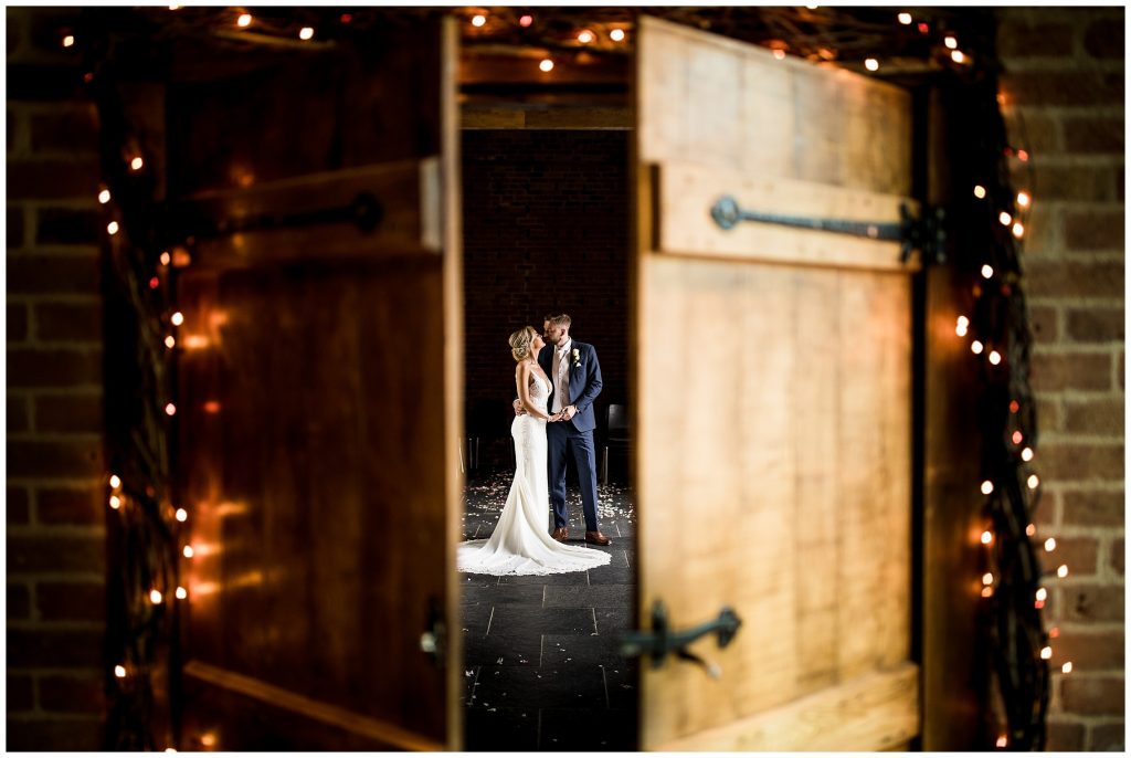 bride and groom kissing through slightly opened doors at redhouse barn, surrounded by warm fairy lights.