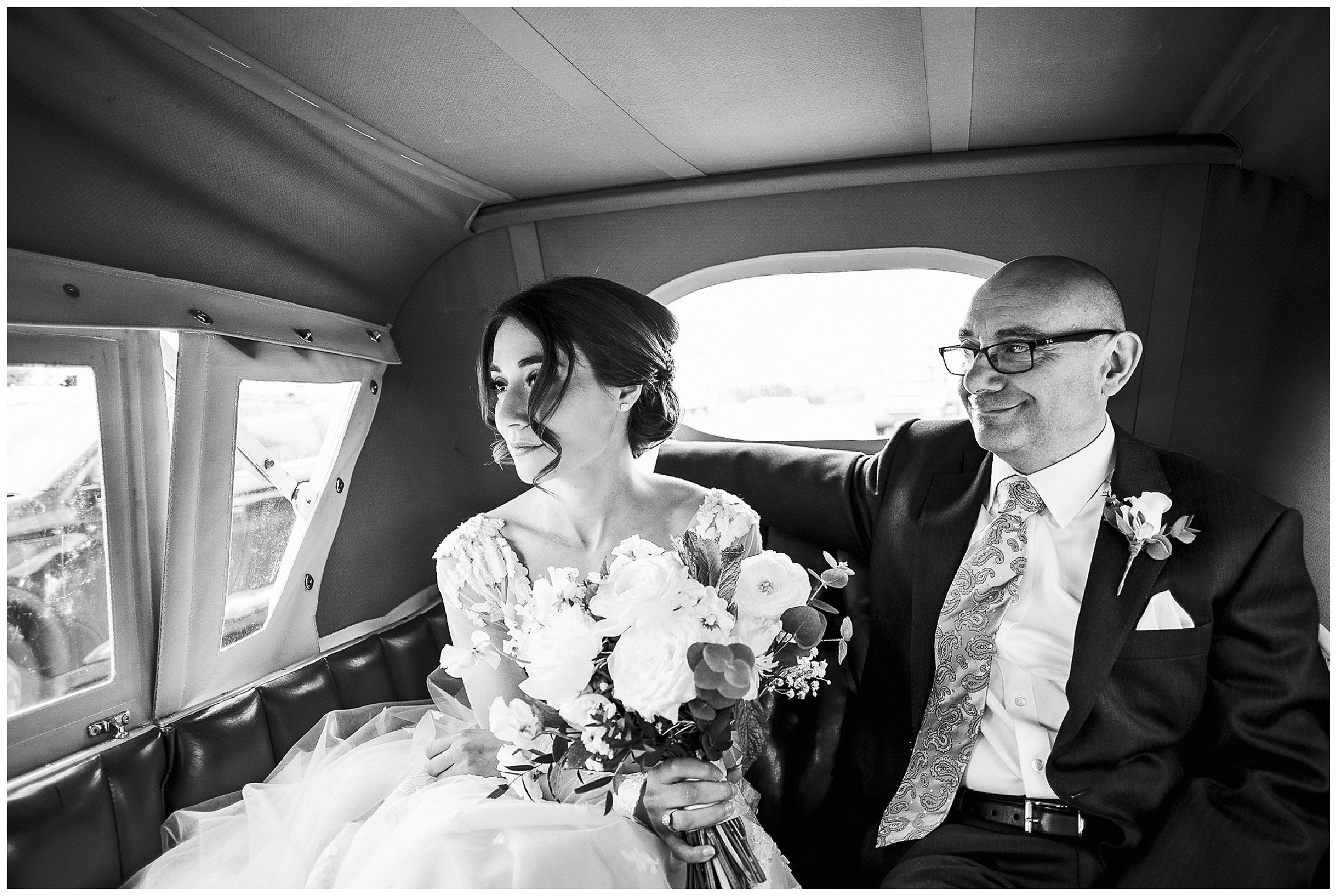 black and white image of bride in wedding car looking out of window as father of the bride looks at her and smiles