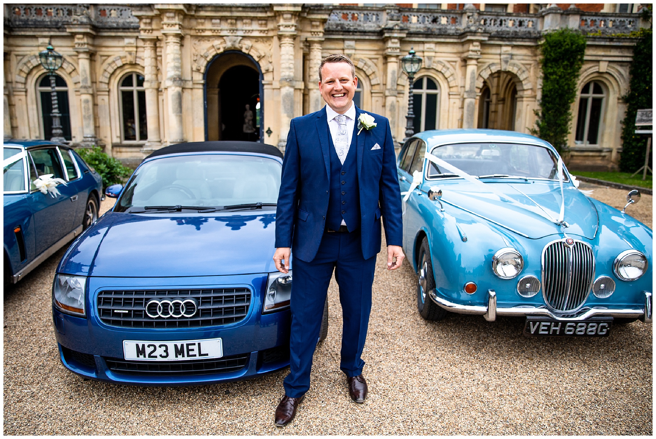 groom in blue suit smiling stood between two blue cars