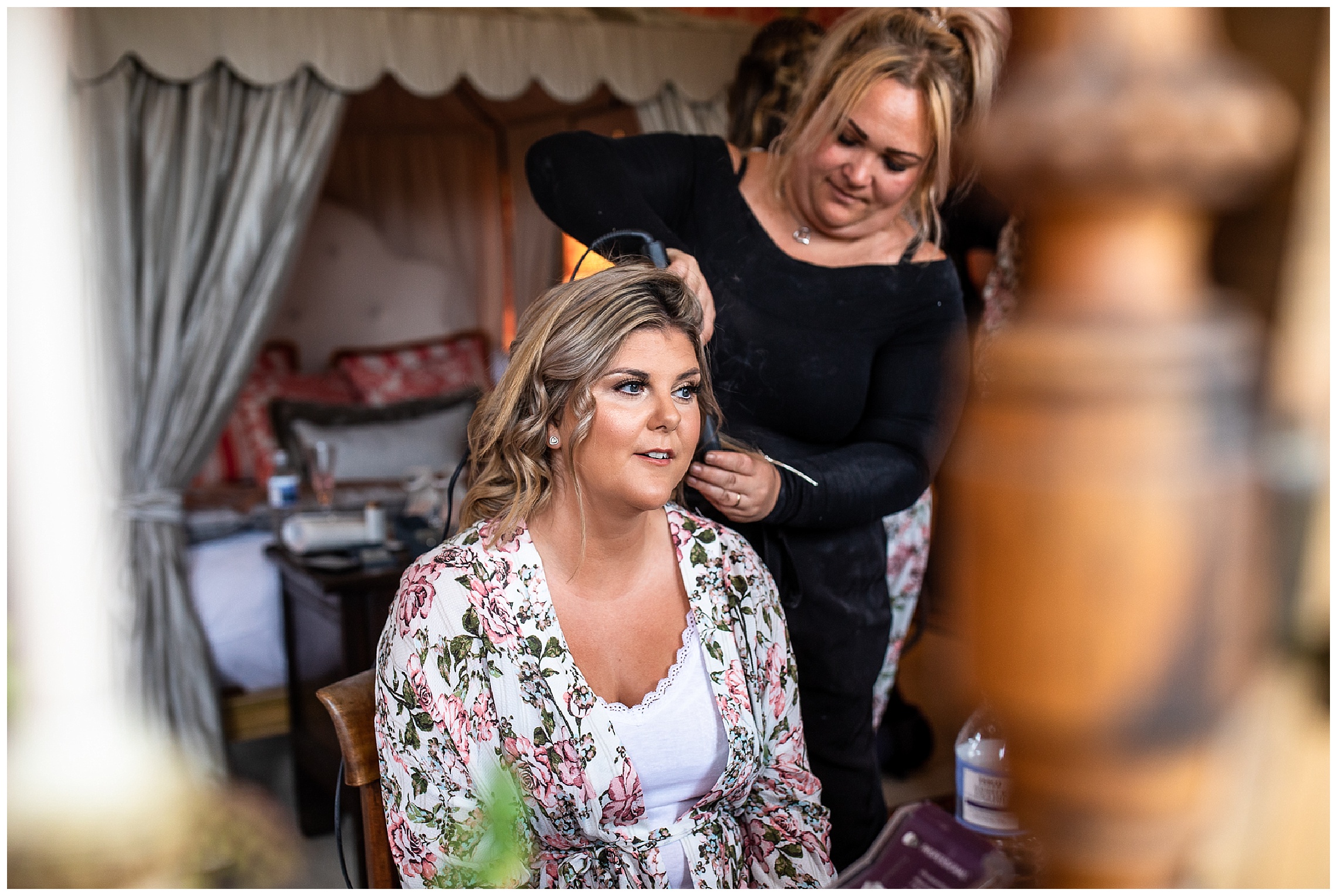 bride in floral dressing gown in pink dressing room having hair curled by stylist using curling iron