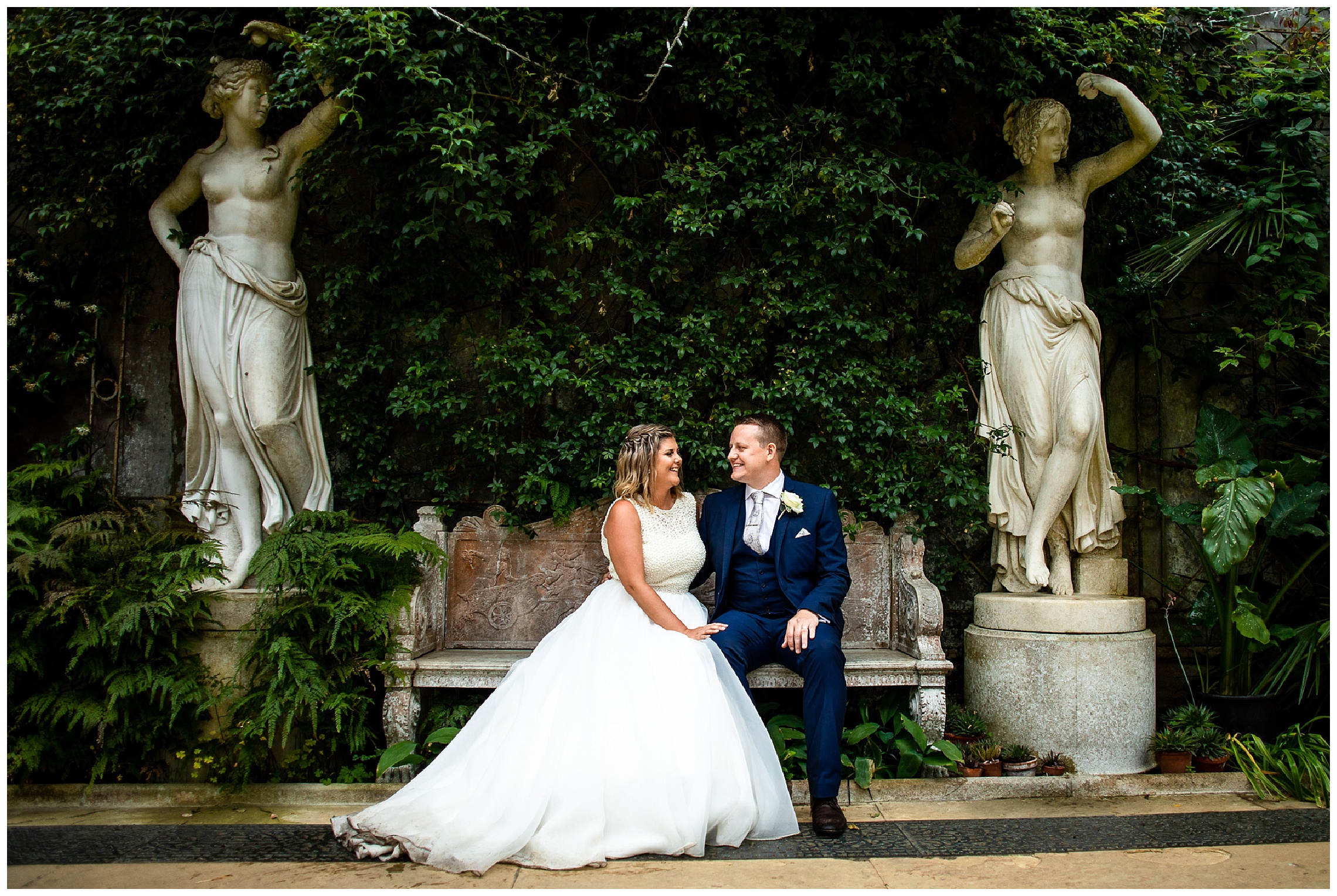 bride and groom sit on marble bench next to statues