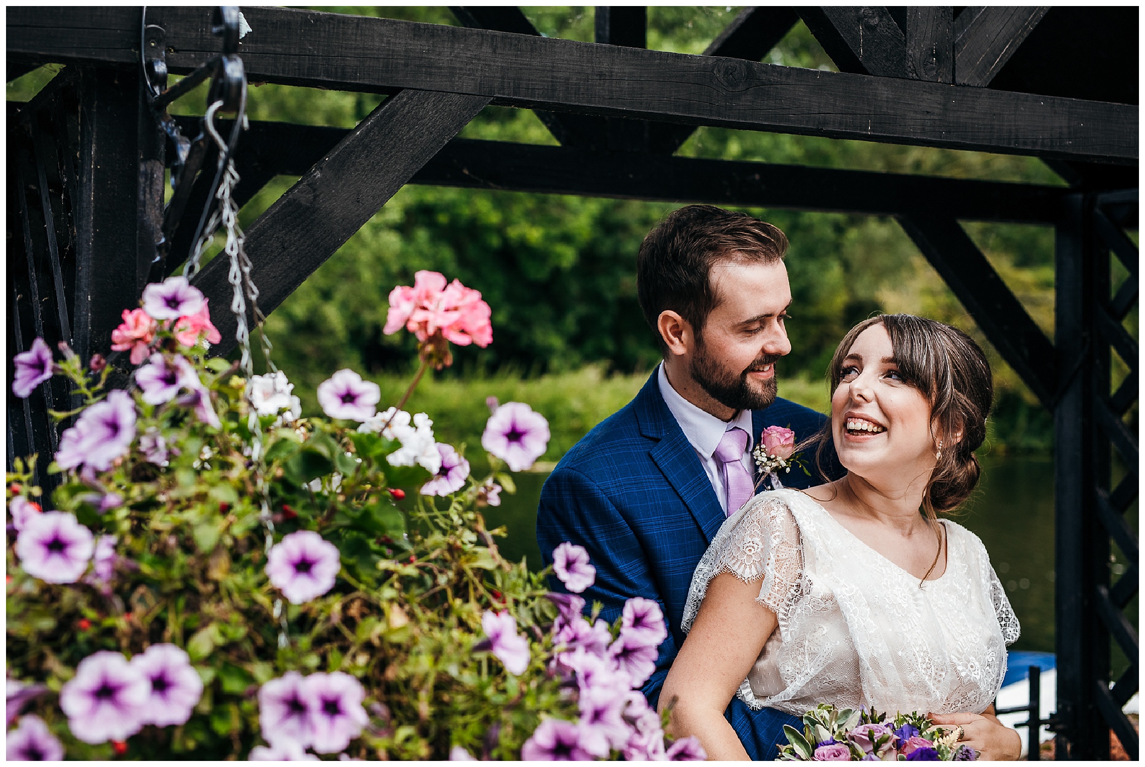 Groom in blue suit standing behind bride in vintage lace dress holding onto her pregnancy bump and looking at her smiling