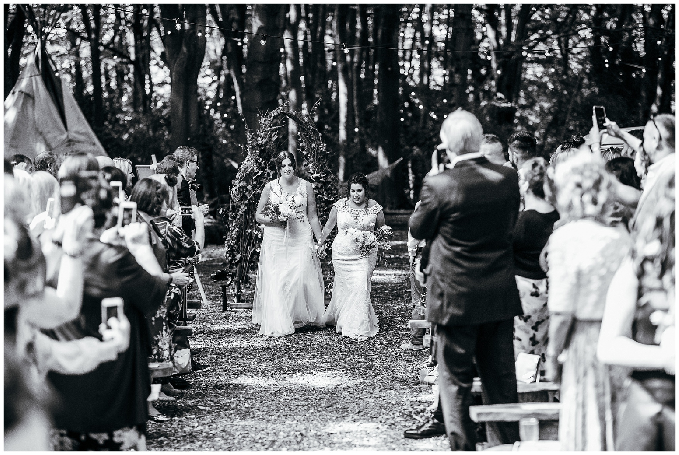 black and white image of brides walking hand in hand down aisle at woodland weddings lilas wood wedding venue