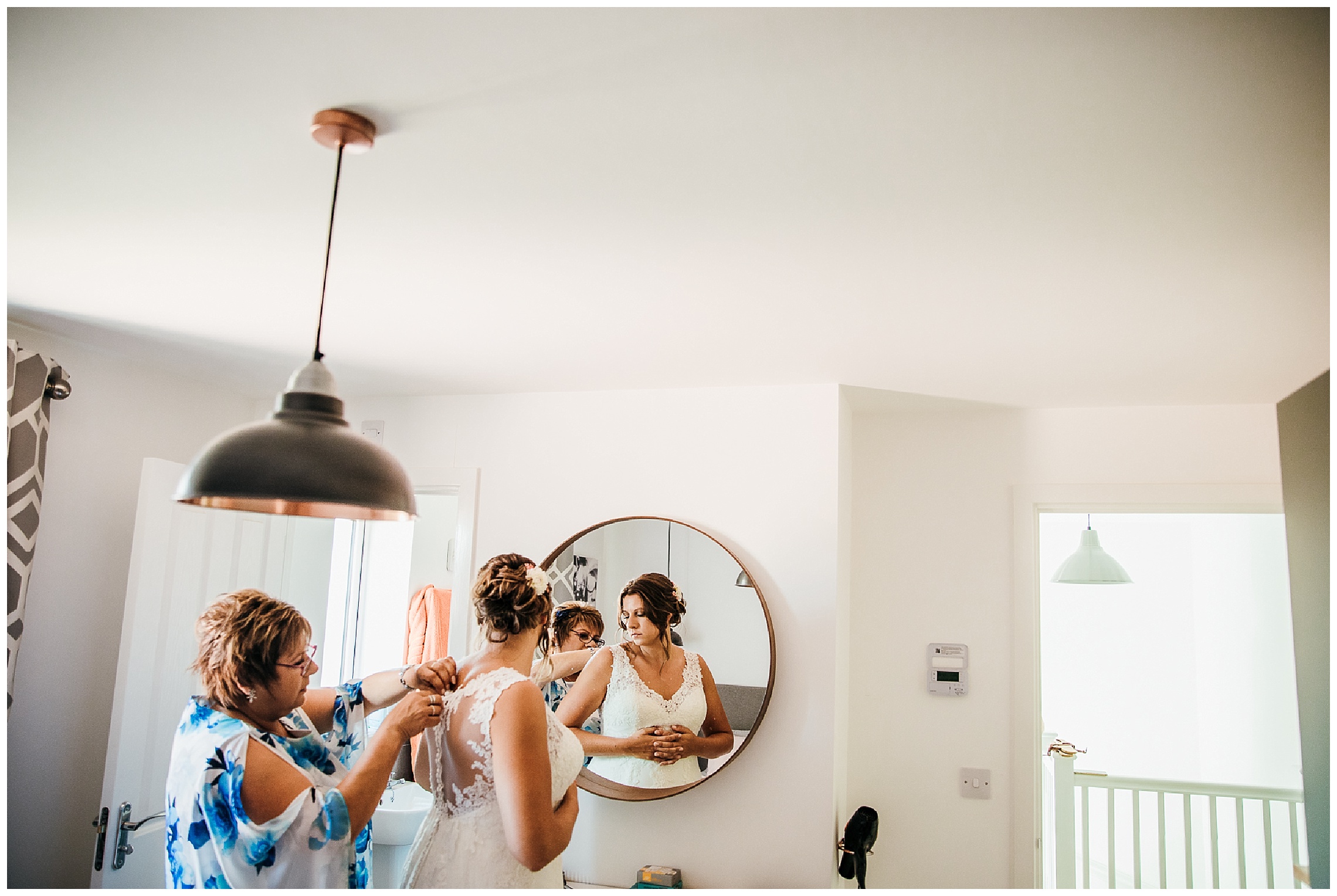 bride having her dress adjusted by her mum standing in white bedroom in front of mirror