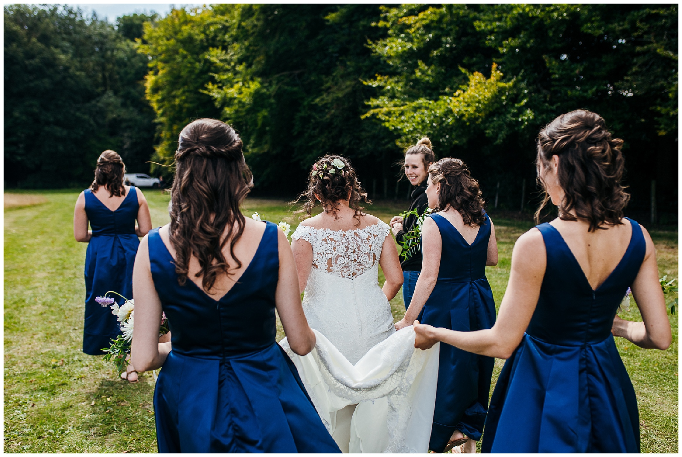 bride walking through meadow with bridesmaids holding her dress off the floor