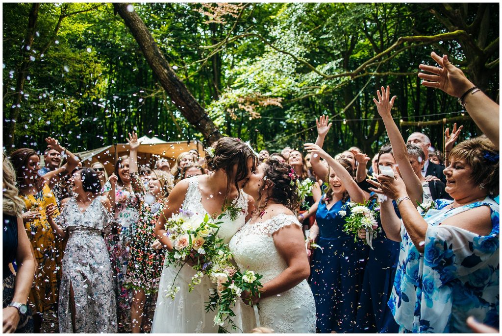 brides kiss in white dresses as guests throw confetti on them