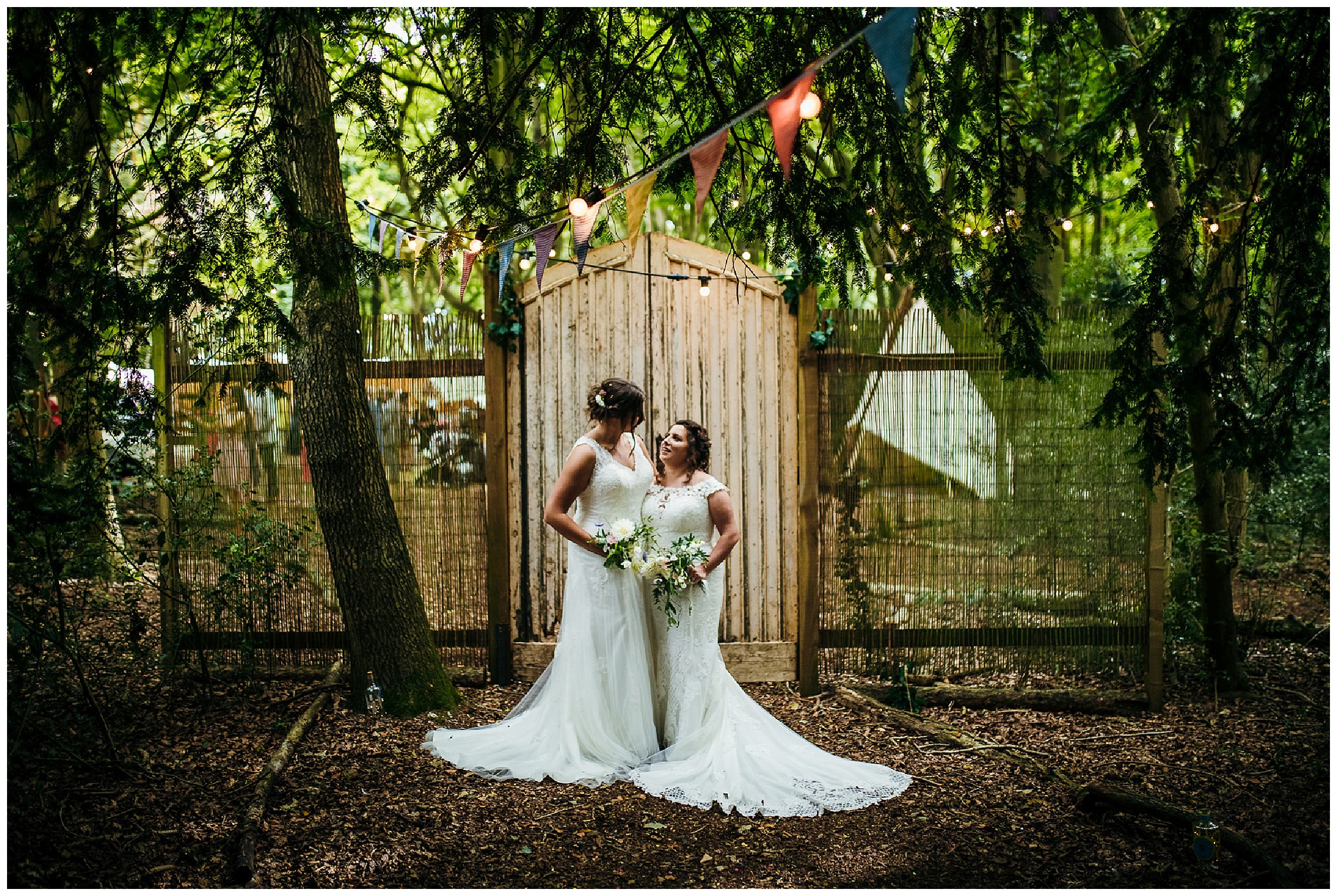 brides in white dresses stand together under bunting at woodland wedding venue