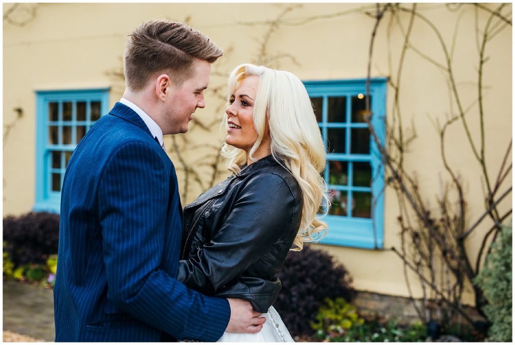 bride and groom standing face to face in front of blue window frames and yellow house at south farm