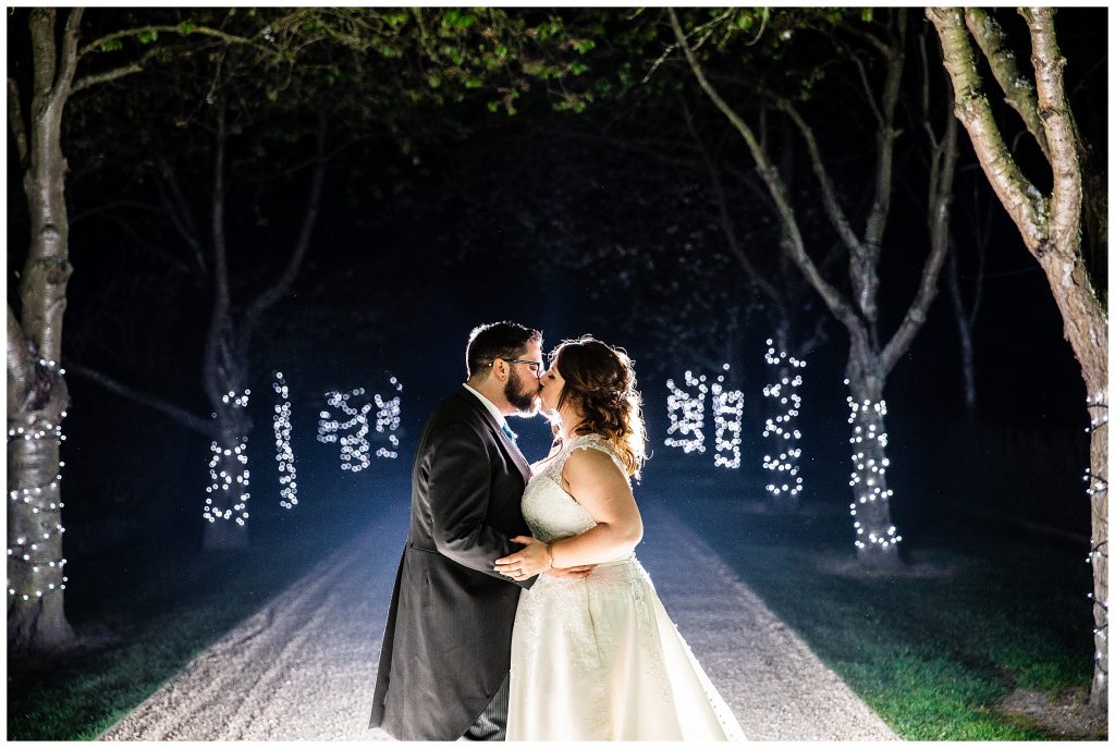 bride and groom kissing in dark driveway dimly lit by lots of fairy lights on trees at south farm