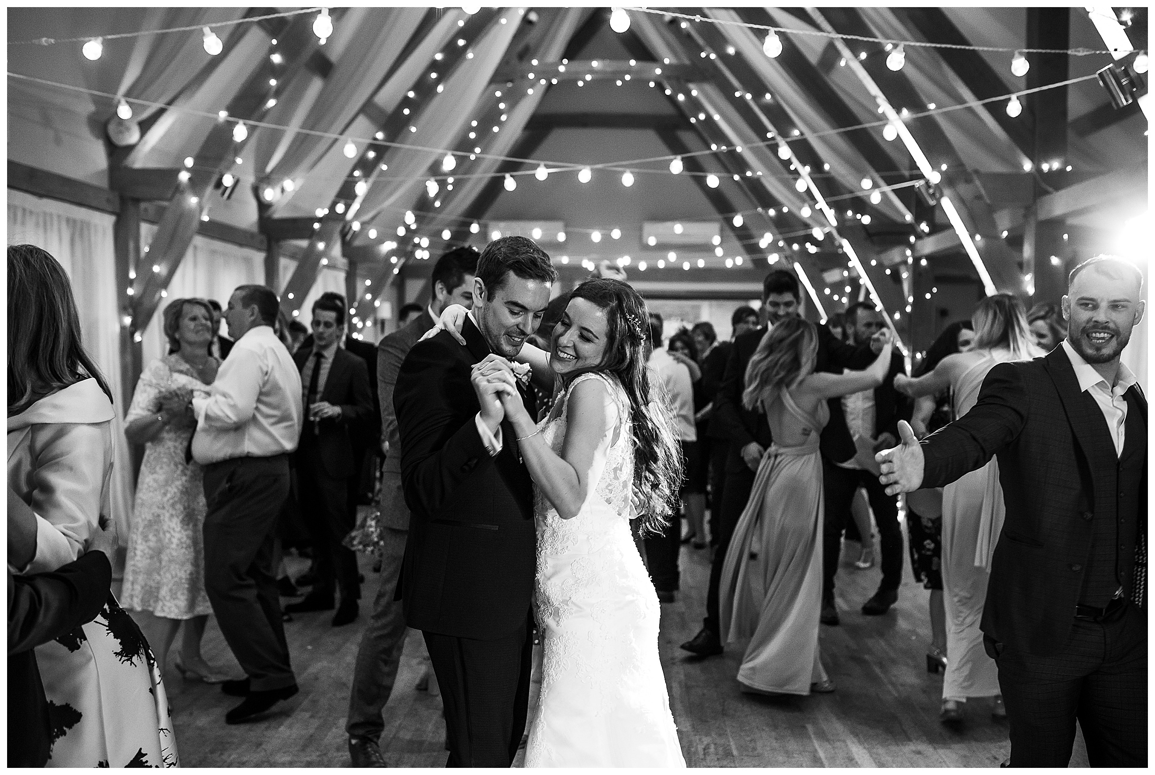 black and white image of bride and groom embracing during first dance surrounded by dancing guests at bassmead manor barns