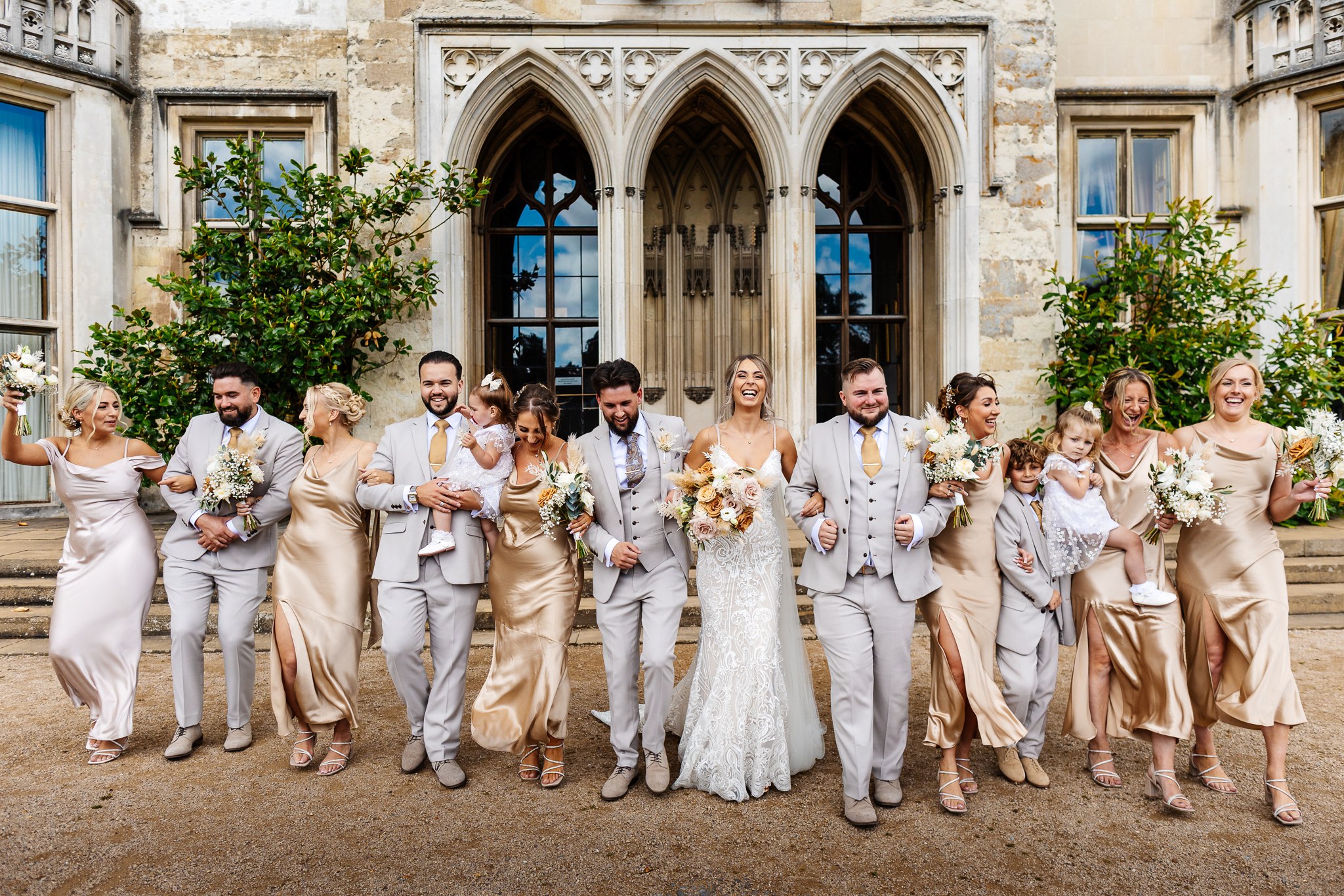 bridesmaids in champagne coloured dresses and gromsmen in light suits with gold ties, with bride and groom at ashridge house, laughing and walking together 