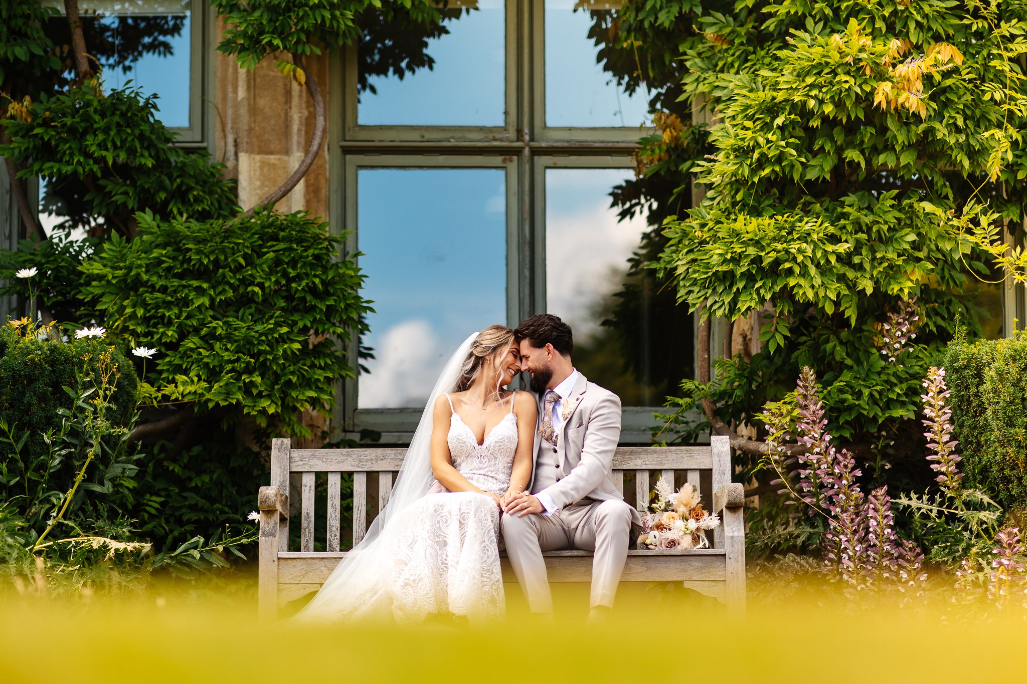 bride and groom sitting together closely on bench in gardens at ashridge house, surrounded by wisteria plants 