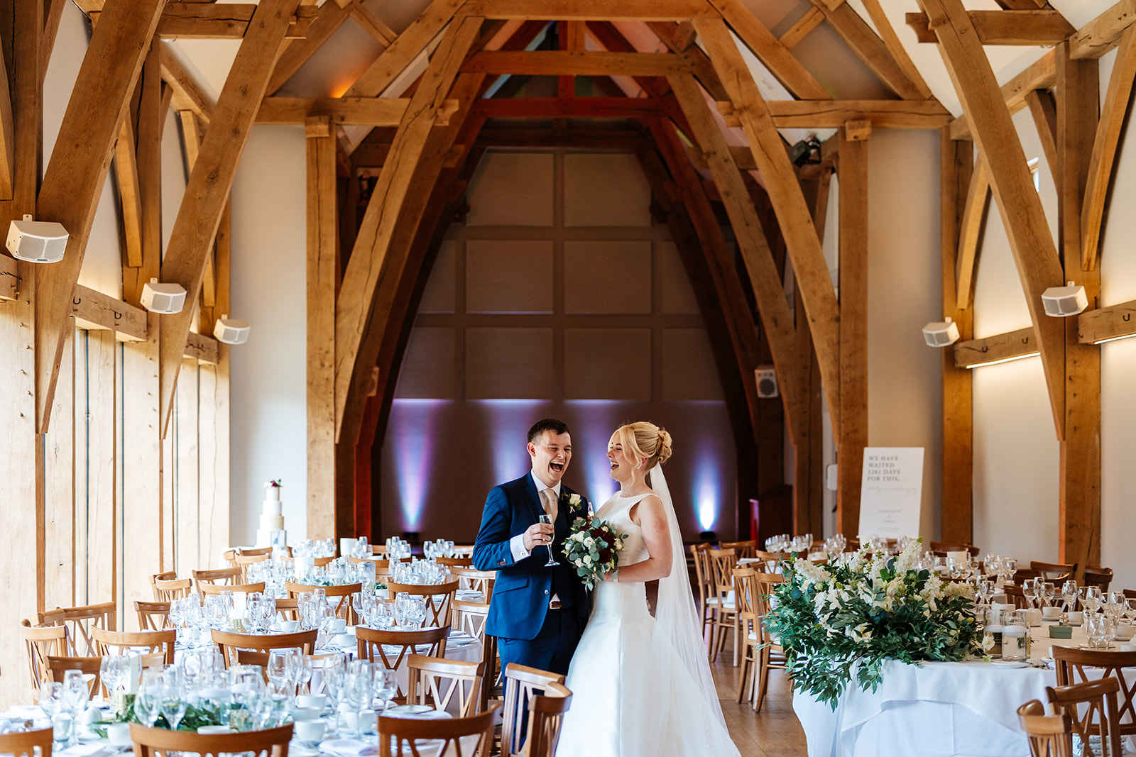 bride and groom stood in the middle of wedding breakfast reception room at Mill Barns wedding venue. They're holding drinks and laughing together.