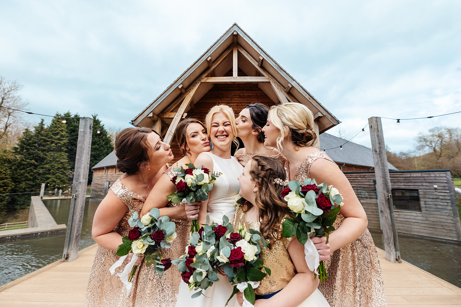 bridesmaids in gold dresses kissing bride, theyre all holding onto red and white flowers 