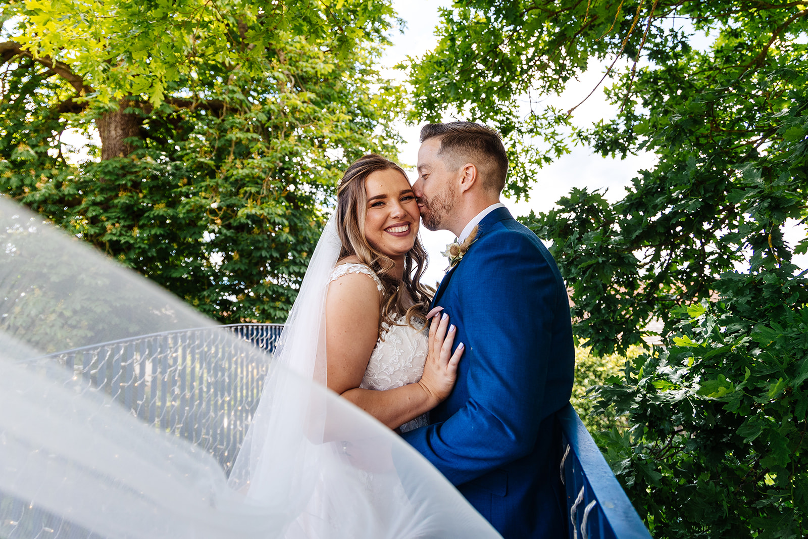 Groom kisses bride on the cheek, her veil blows in the wind