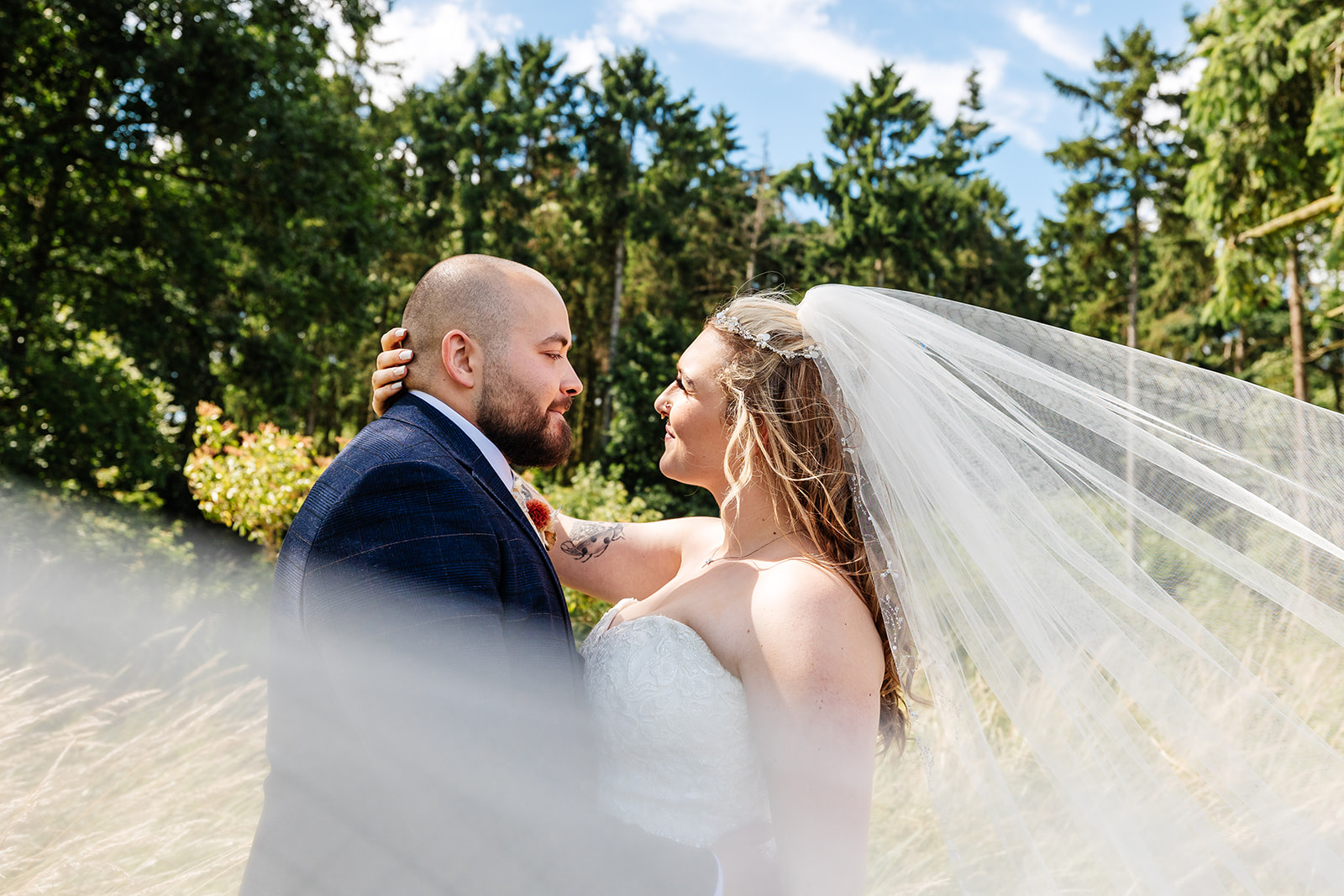 Couple are looking into each others eyes, brides veil is draping across them. Outside in wooded area 