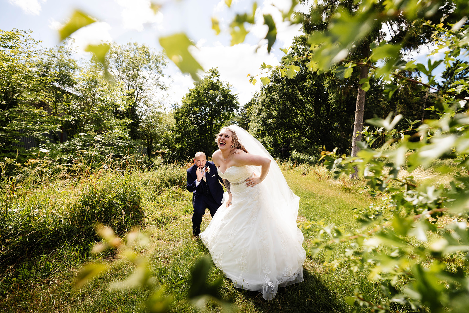 Couple walking through wooded area, groom is posing and the bride is laughing 