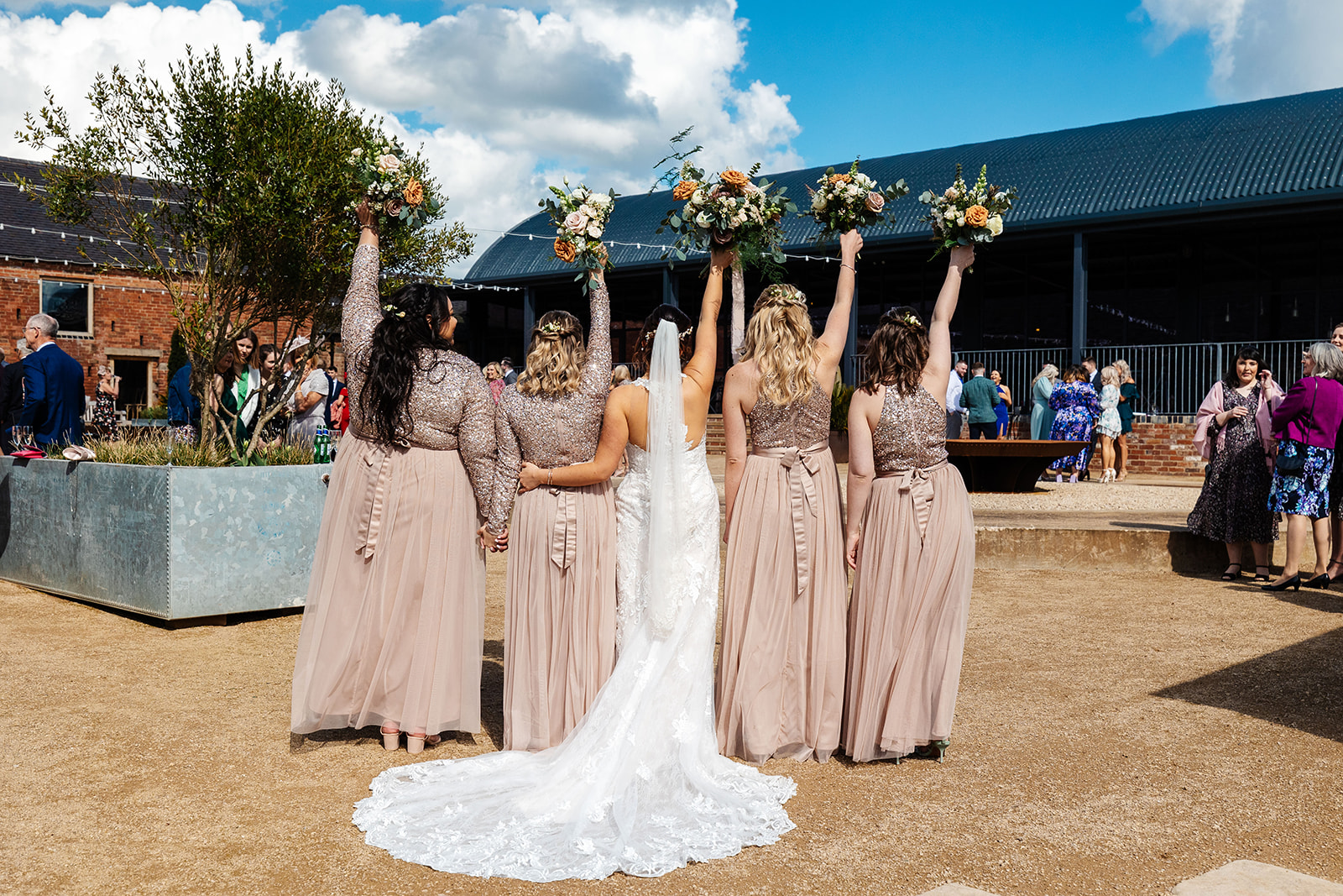 Bride and bridesmaids in pink sequin topped dresses with backs to camera holding bouquets up  