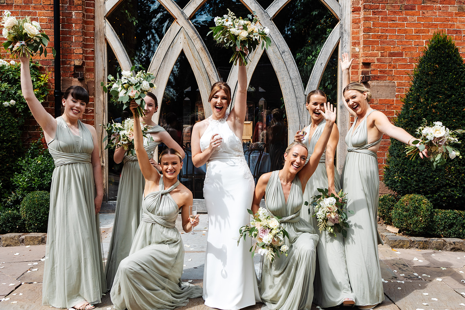 Bride and bridesmaids (in sage green dresses) holding up bouquets and laughing outside ceremony space
