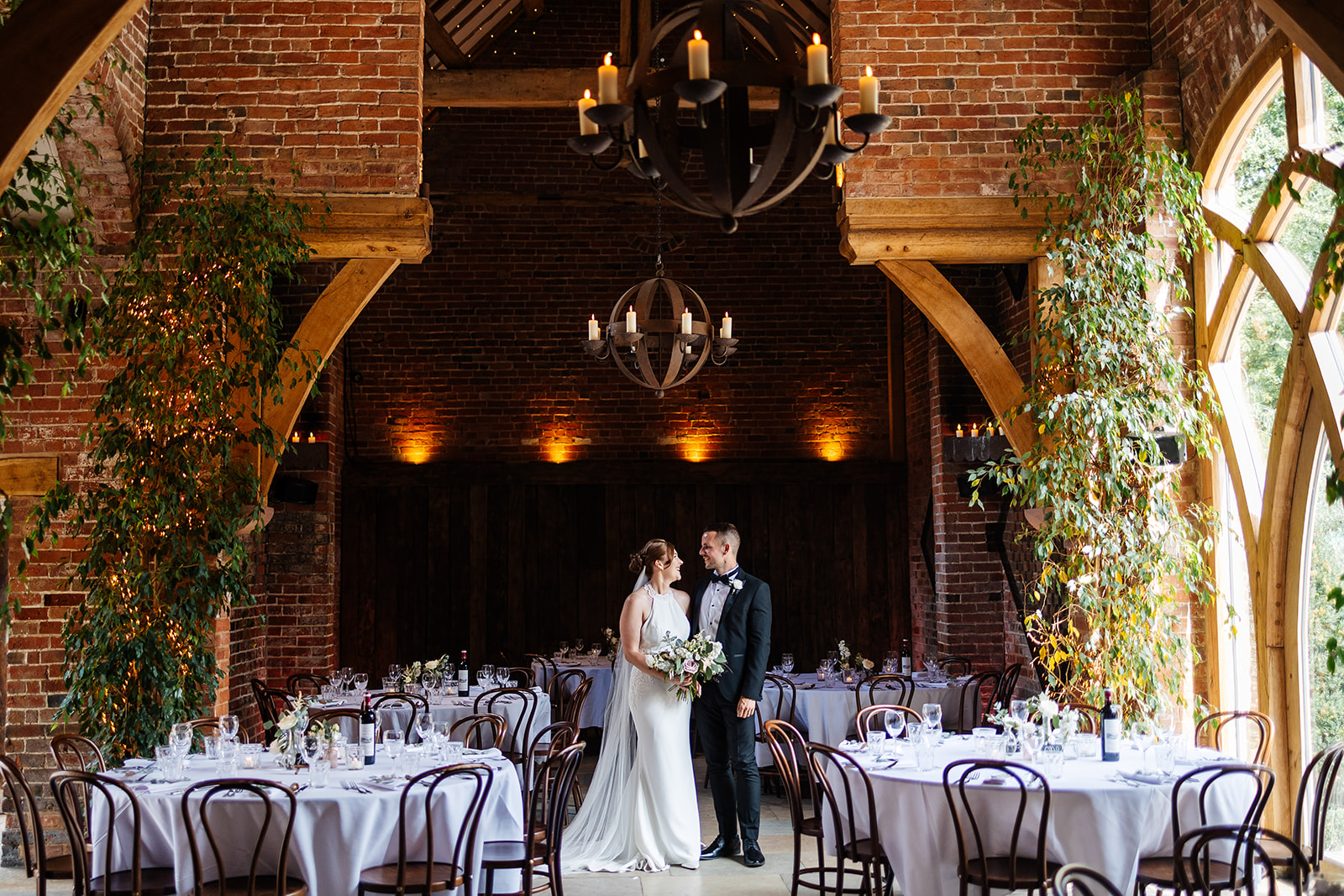 Couple in space set up for wedding breakfast. The inside has exposed brickwork, tall foliage and light pouring in from a tall window 