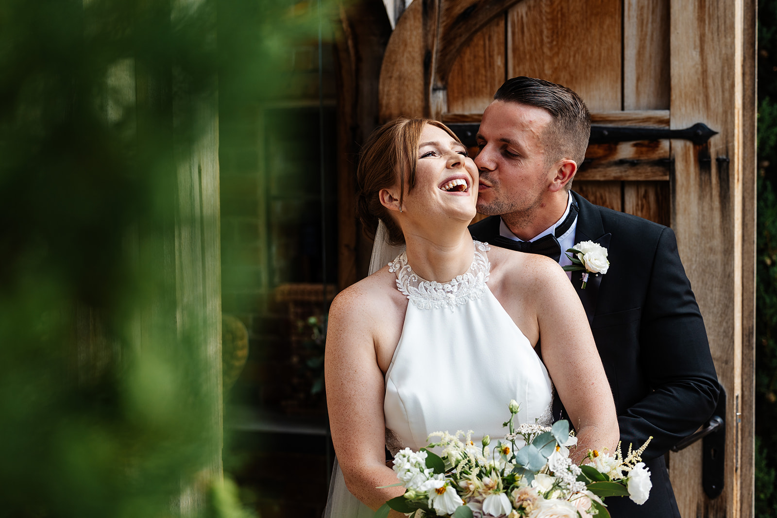 Groom gives bride a kiss on the cheek, they are outside in front of a large wooden door, she is smiling 