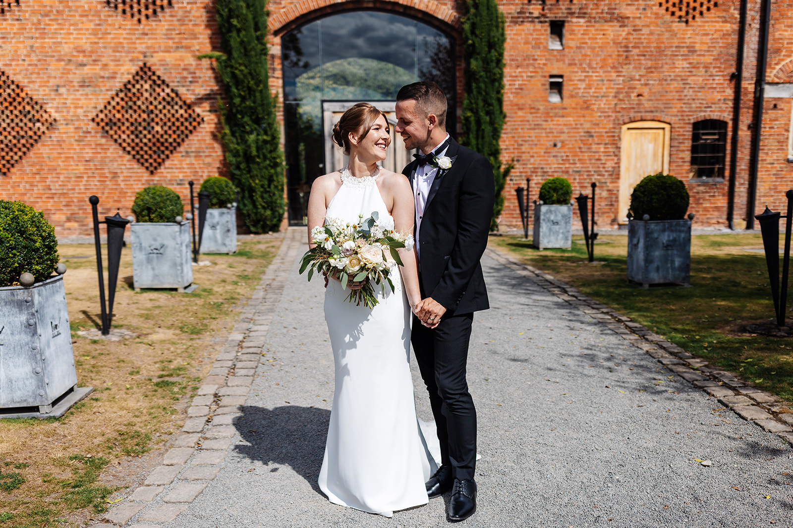 Couple stand and hold hands, the walkway to Shustoke Barn and the large glass window is in the backgound. The sun is shining 