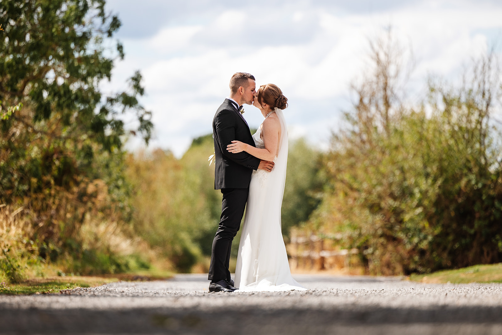 Couple share a kiss on drive way to  Shustoke Barn, trees and greenery are in the background on either side 