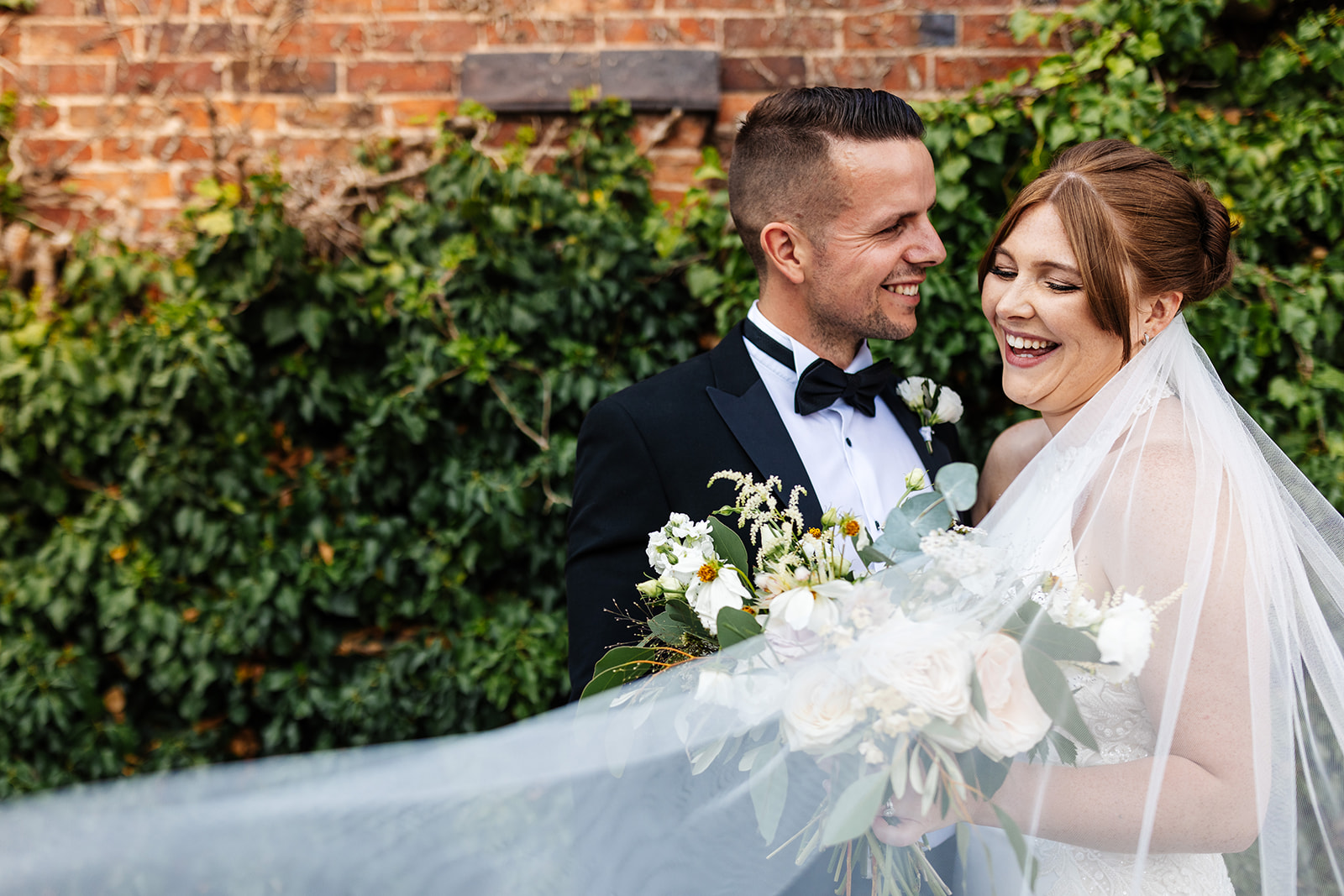 Couple are outside laughing, brides veil sweeps across them 