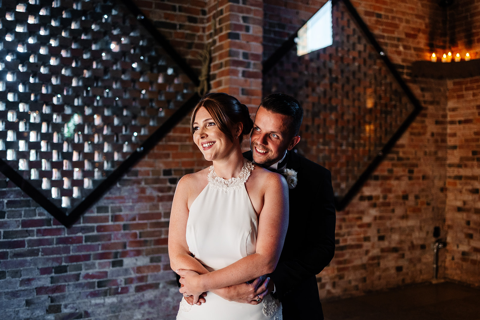 Couple are looking into the distance, groom has his arms around bride, exposed brickwork is in the background 