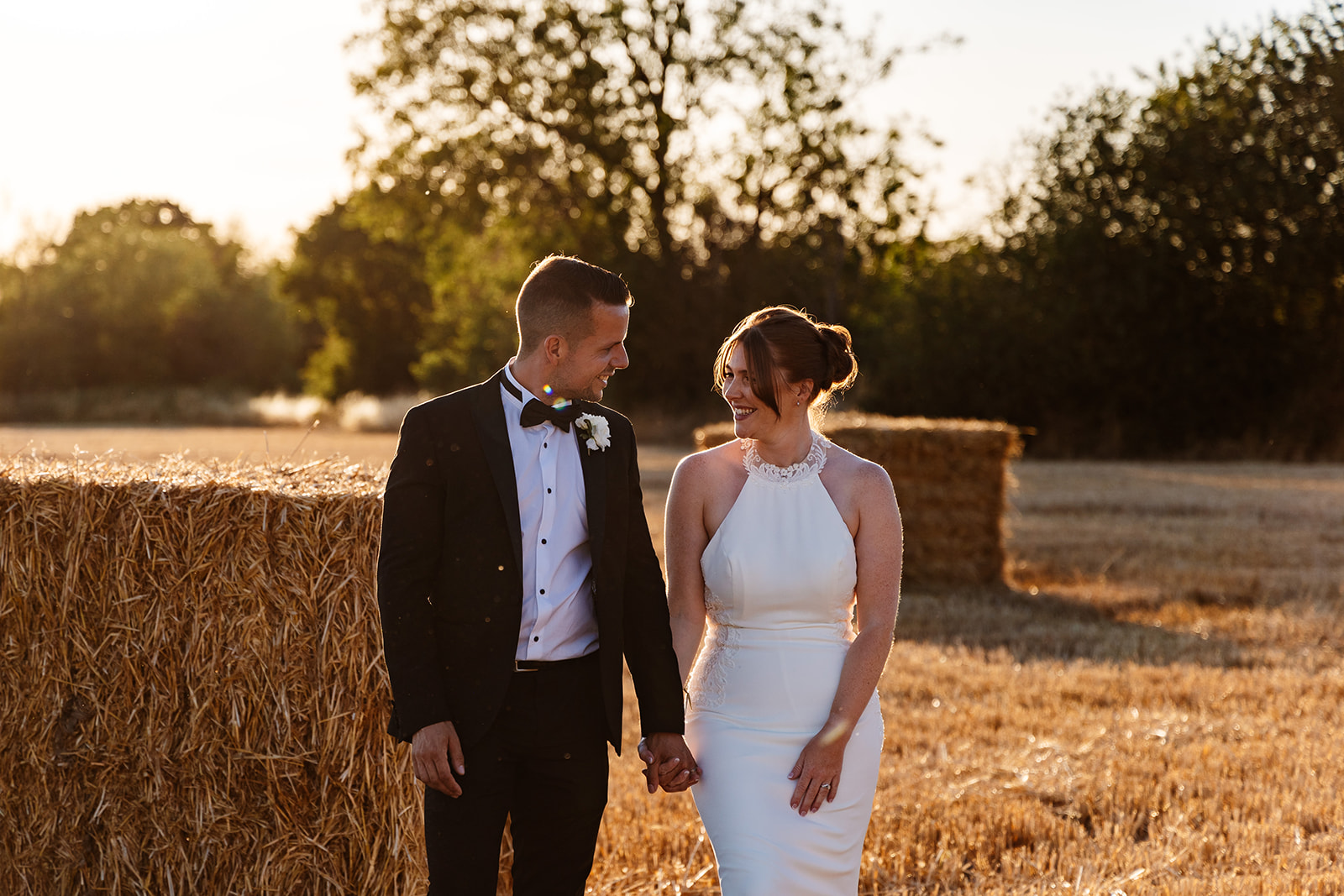 Couple take a walk at sunset in a field with trees and hay bales, smiling at each other. 