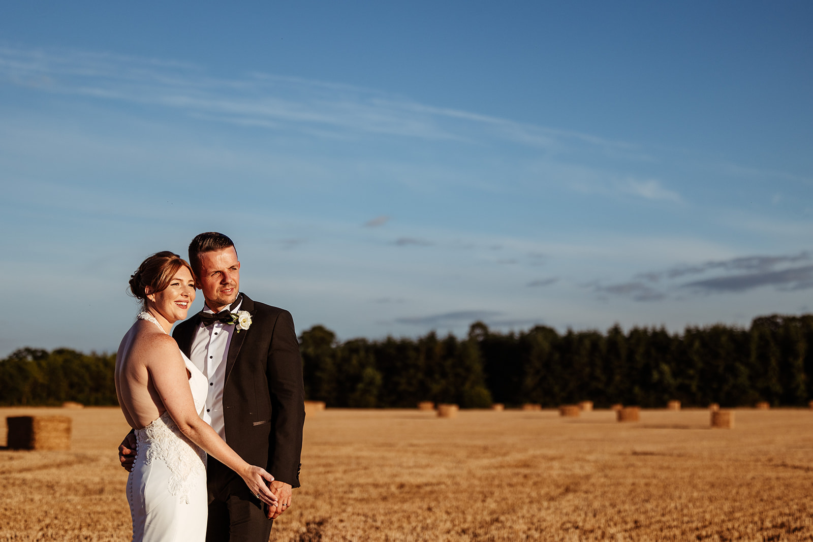 Couple watch the sunset in a field with trees and hay bales, holding hands 