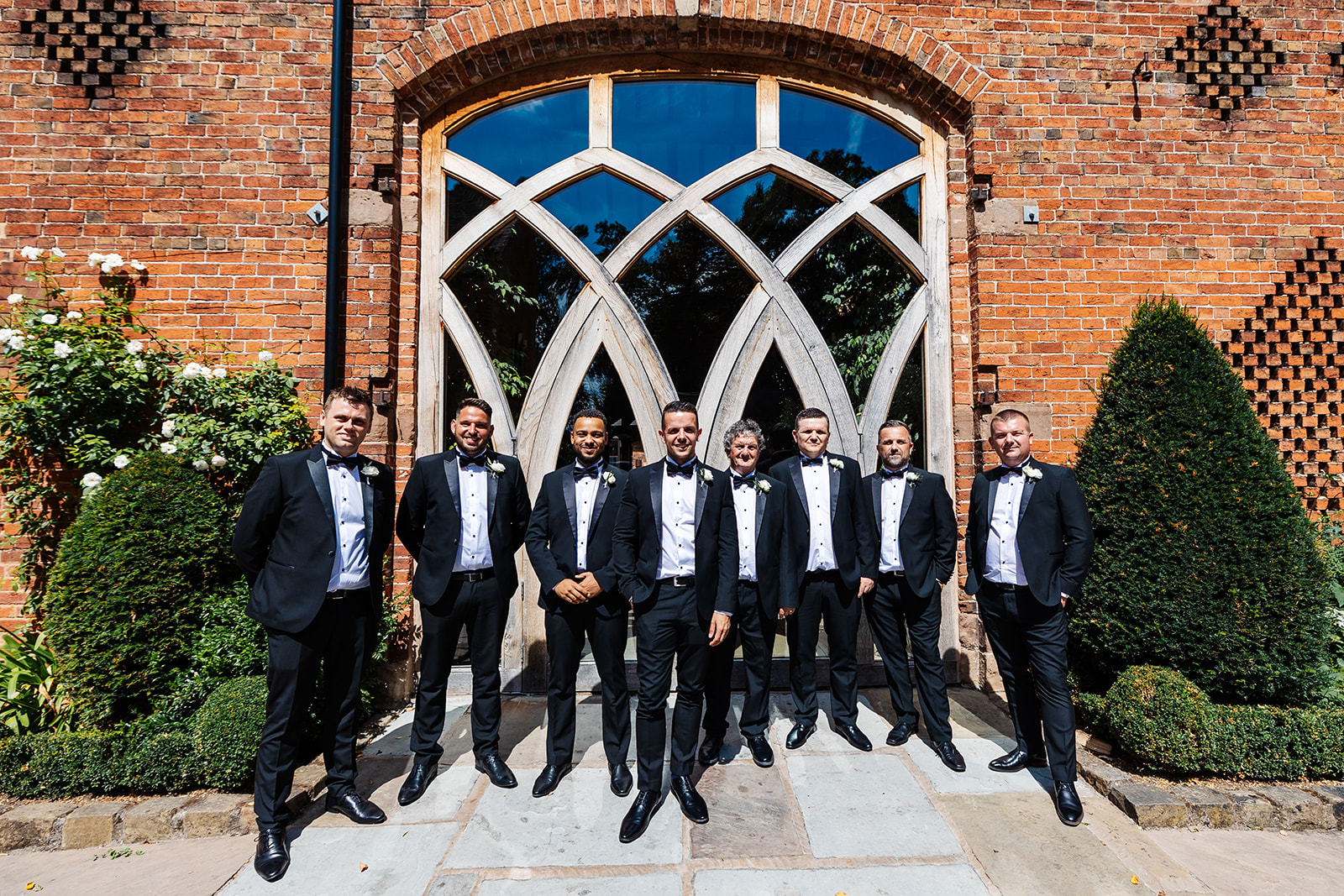 Groomsmen outside Shustoke Bar all in dark suits with bowties, the sun is shining 