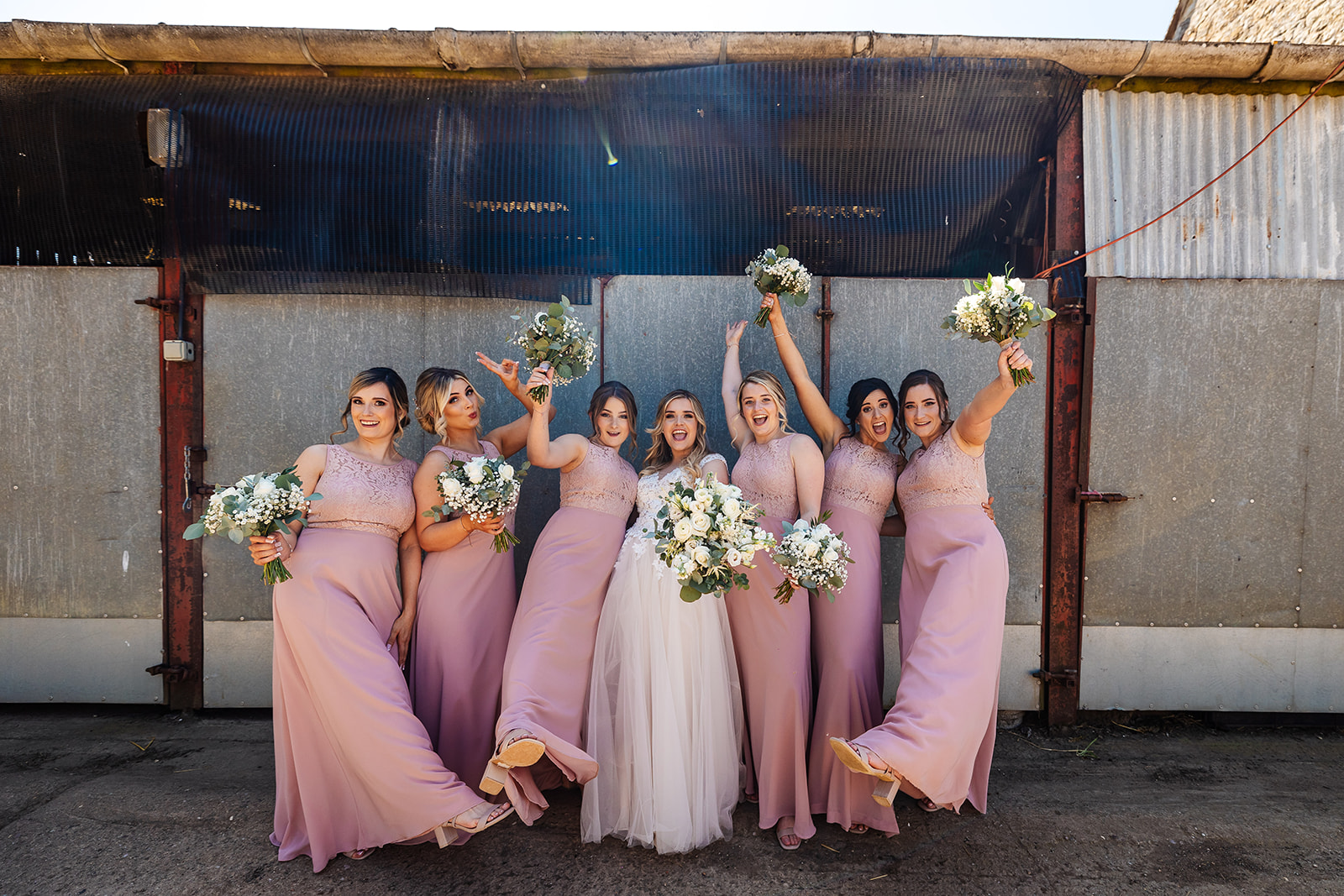 Bride with bridesmaids outside metal barn holding up bouquets 