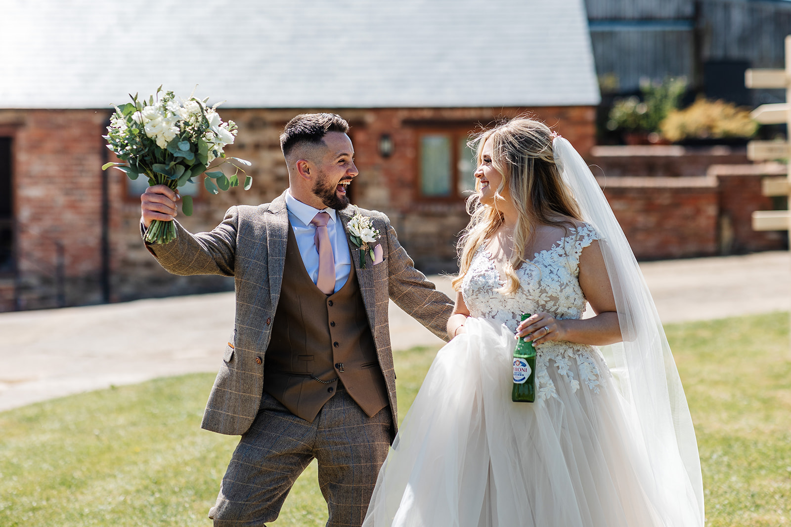 Couple walking, bride has a bottle of beer in her hand with the groom holding up her bouquet 